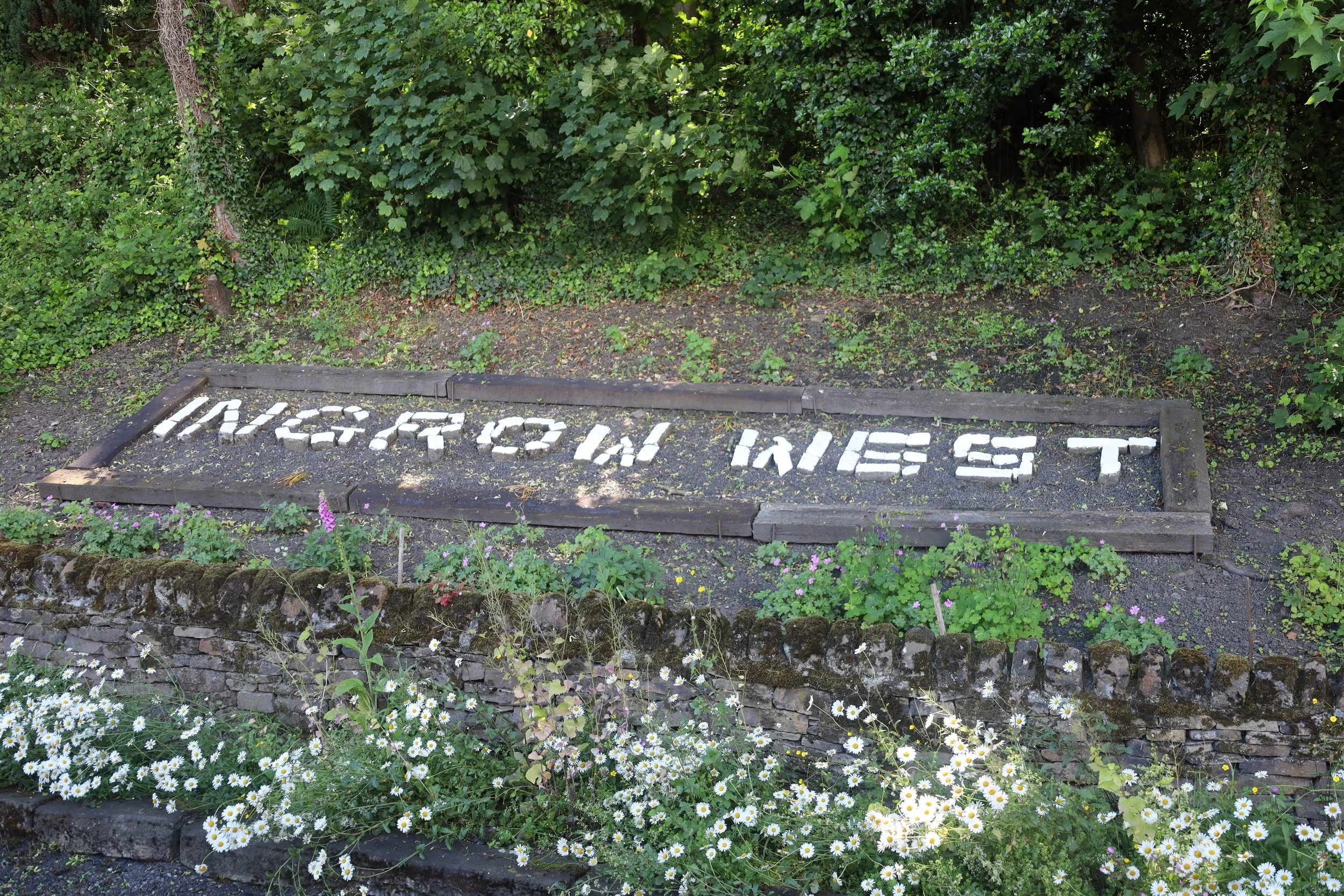A garden with a raised flower bed made of wooden planks. Inside the flower bed, white stones spell out 'LOVE YOU ST'. The garden is lush with various green plants and shrubs, with small white and purple flowers blooming around the edges.