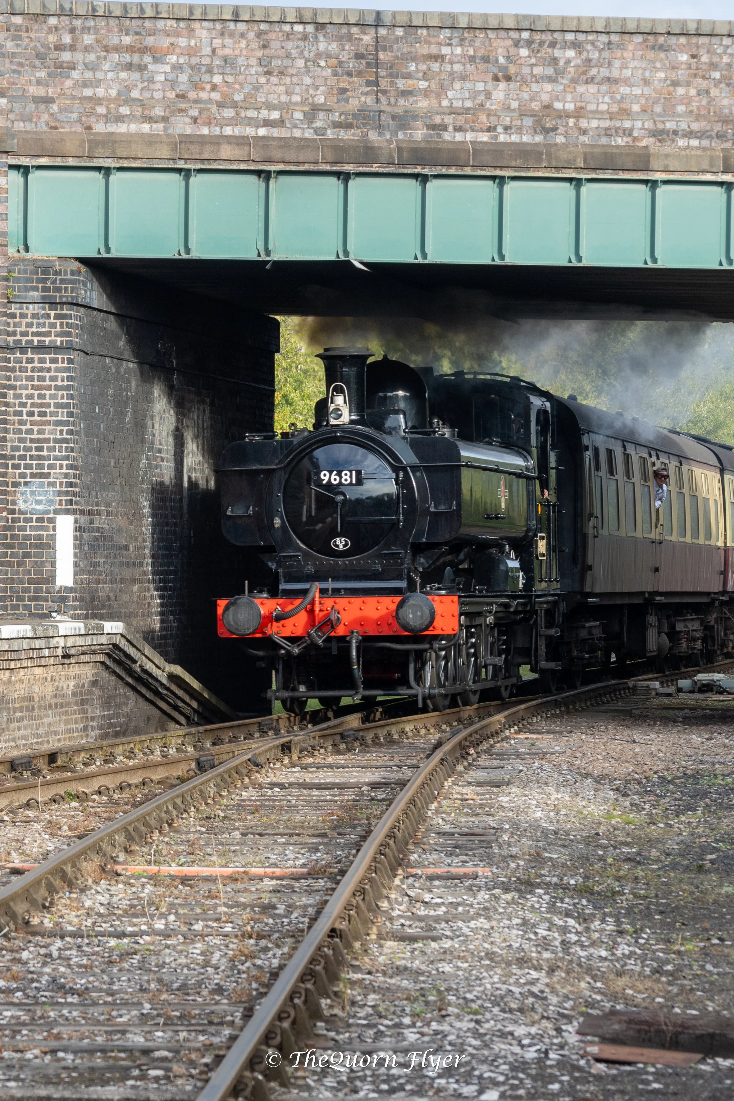 A vintage black steam locomotive train passing under a brick and metal bridge, with steam rising from the engine and the train moving along railway tracks.