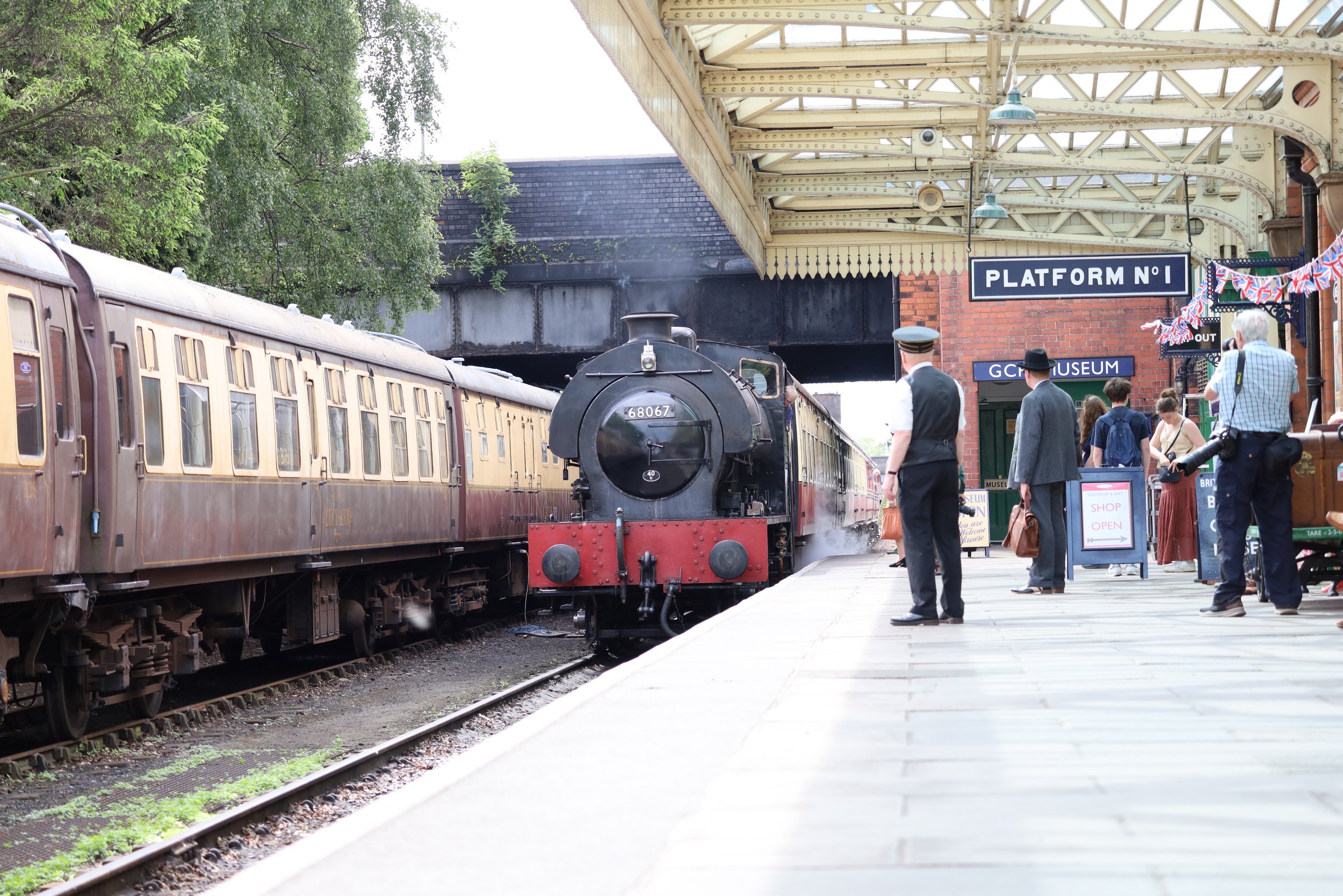 A vintage train at a station with people waiting on the platform. The train is black and cream-colored and steam is visible coming from its chimney. The station has a yellow iron canopy, and signboards indicating 'Platform No.1' and 'GCM Museum.' Some old-fashioned clothing is worn by the people, and decorative Union Jack bunting hangs from the station wall.