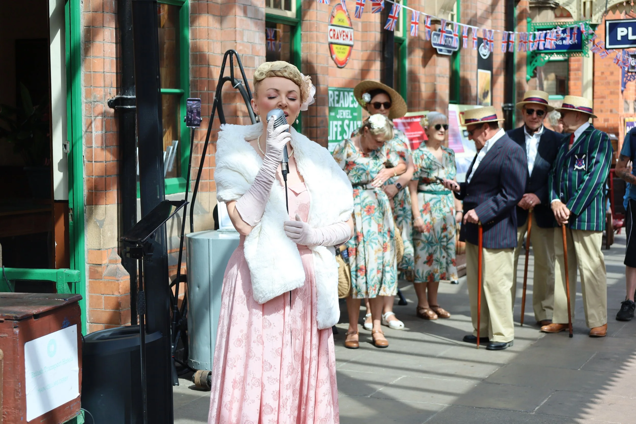 A woman dressed in vintage clothing singing with a microphone on a busy street possibly during a street performance or event. People dressed in retro fashion are gathered around, some wearing hats and sunglasses. The background shows brick buildings and decorative banners.