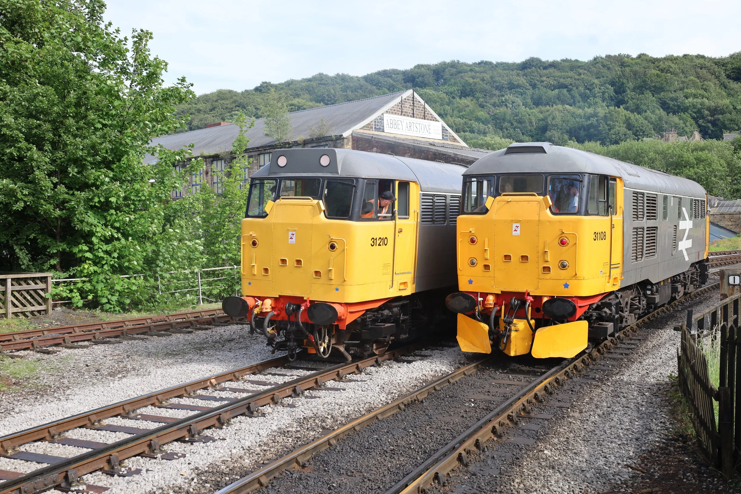 Two yellow and gray diesel locomotives on train tracks surrounded by greenery, with a building labeled 'Abbey Artonne' in the background and hills covered in trees.