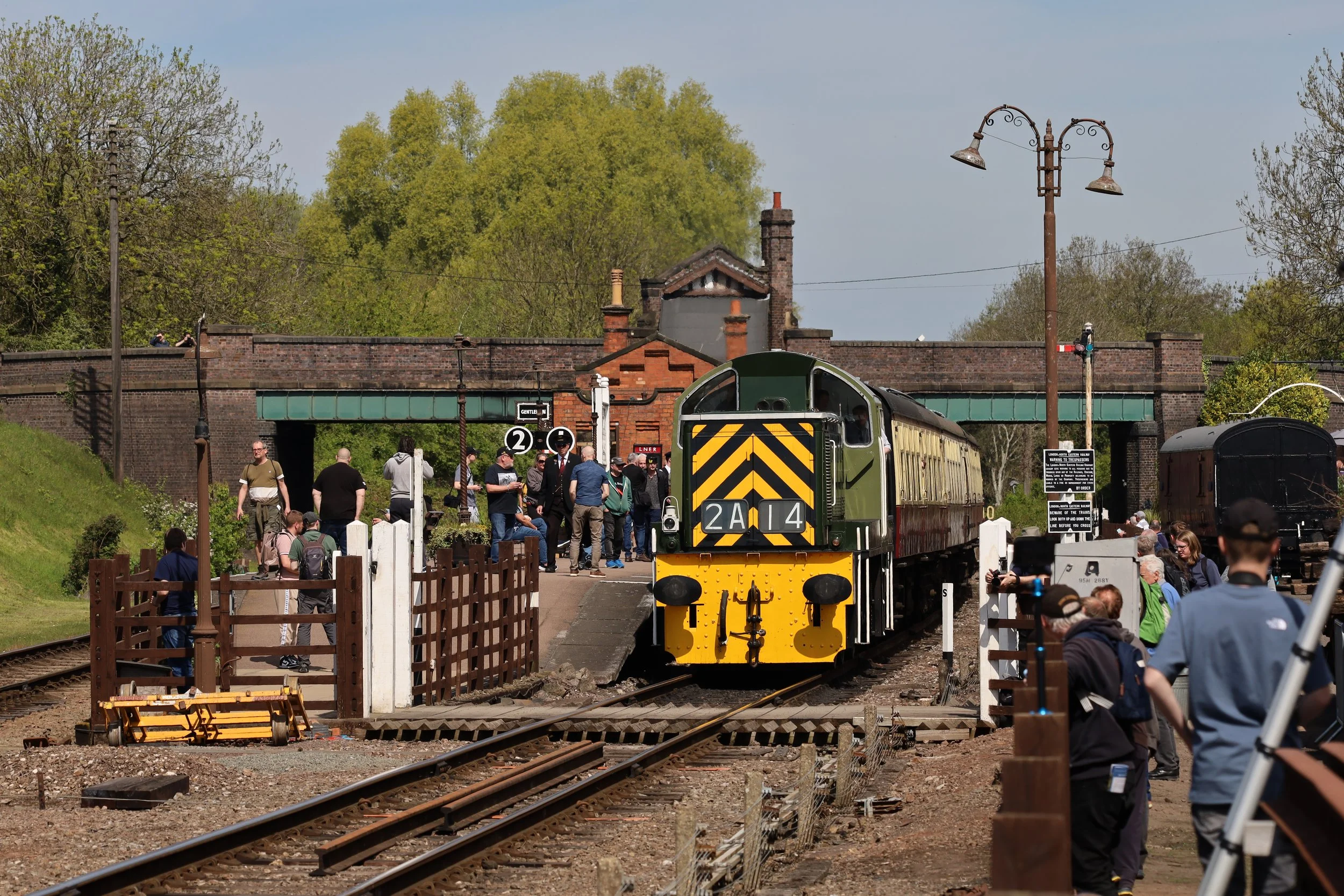 A vintage train arriving at a station with people gathered around, trees in the background, and a brick overpass above, during a sunny day.