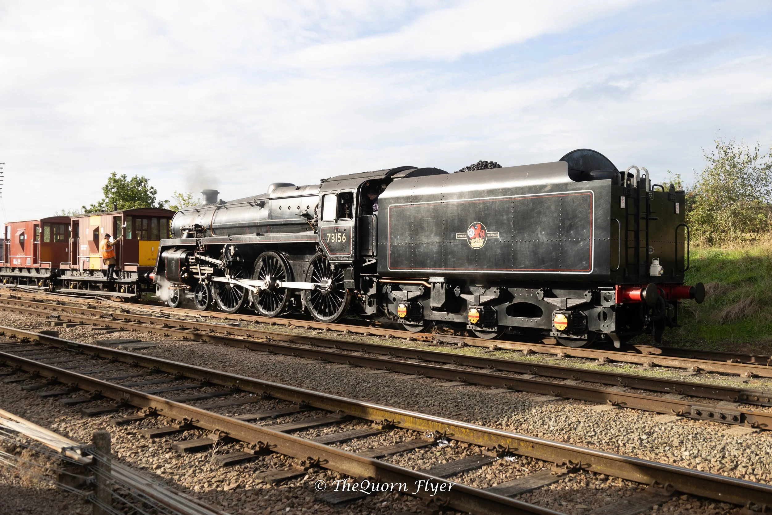 A vintage steam locomotive train with black and silver body, numbered 73156, pulling wooden passenger cars with glass windows on railway tracks outdoors under a partly cloudy sky.