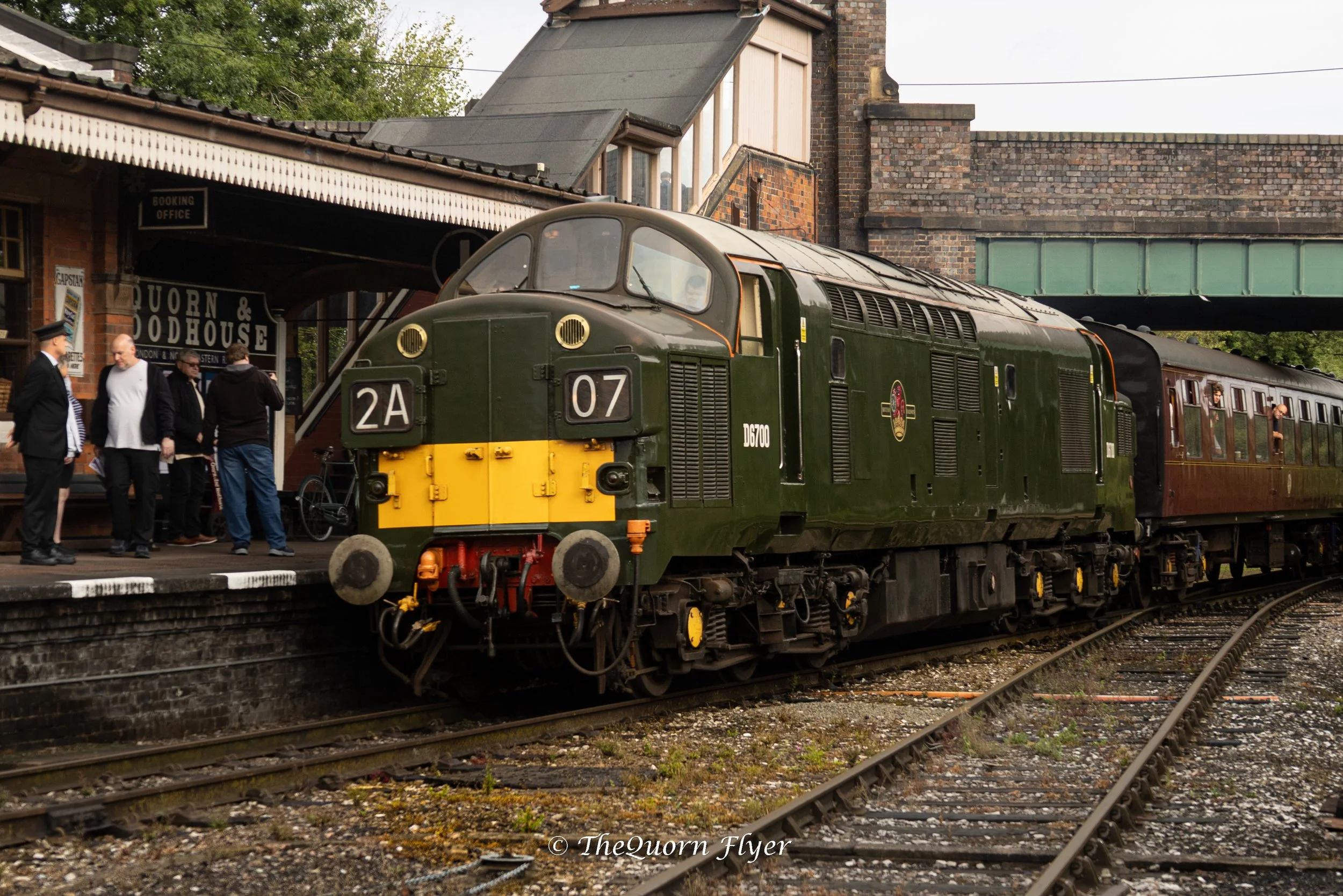 A vintage train pulling into a station with a group of people waiting on the platform. The station building is brick with a sign for the booking office. The train is green with yellow details and old-fashioned passenger cars.