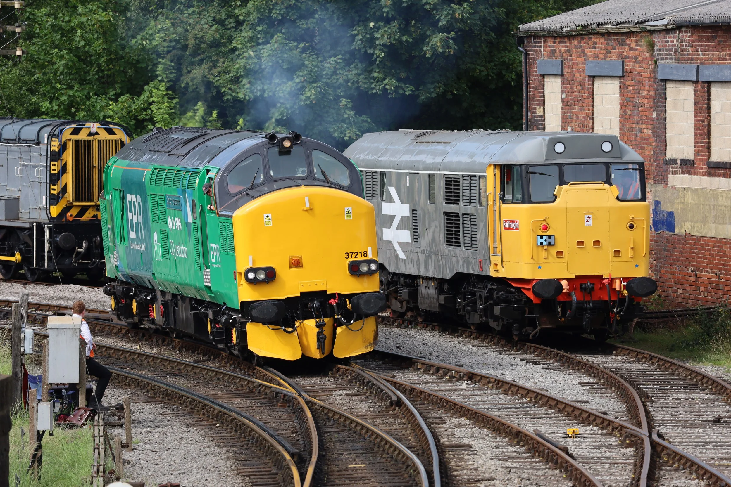 Multiple trains on railway tracks near a brick building, with green trees in the background.