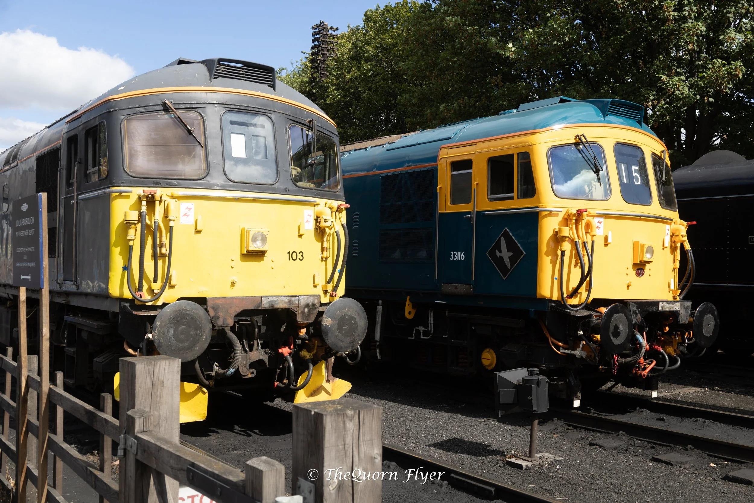 Two vintage train locomotives, one yellow and one blue, parked outdoors next to green trees and a wooden fence.