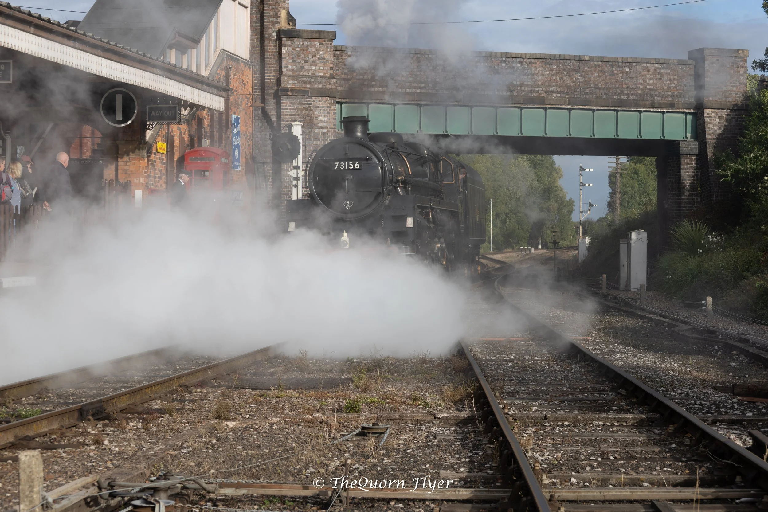 A steam locomotive emerging from a tunnel at a train station, with steam billowing around it and passengers waiting on the platform.