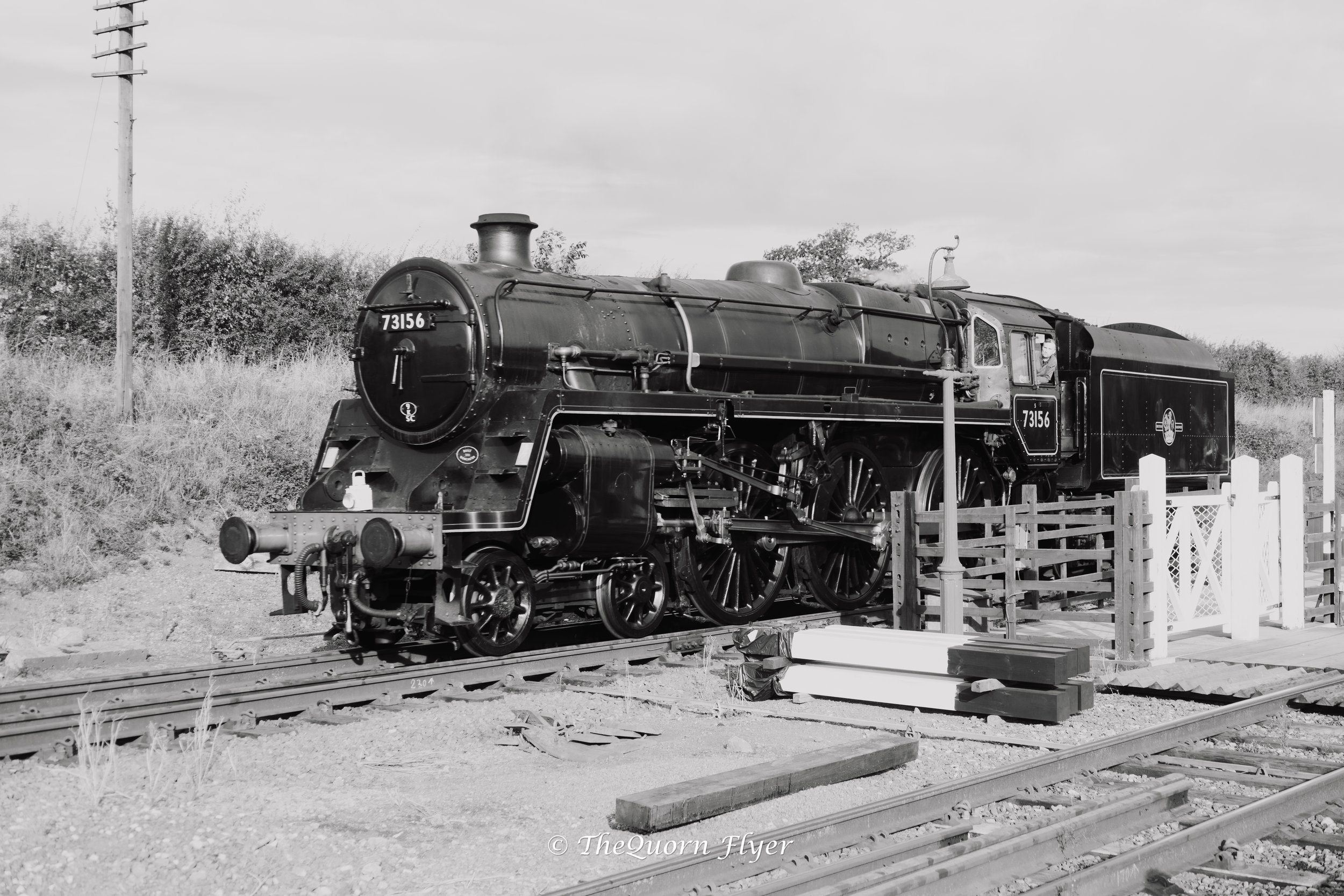 Black and white photo of a vintage steam locomotive with the number 73156, situated on railroad tracks with a wooden fence nearby, and trees and a cloudy sky in the background.