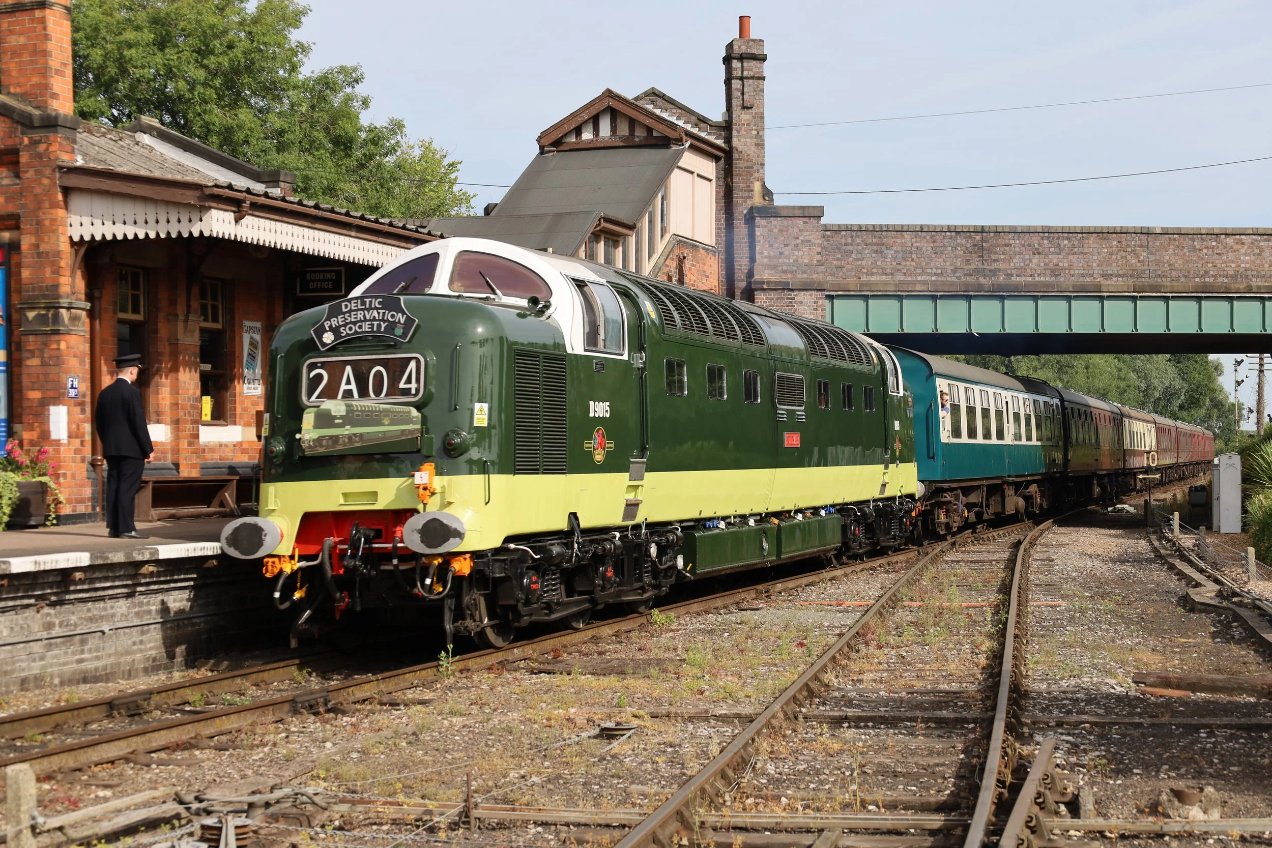 A vintage green railroad locomotive at a train station with a station building and a person in vintage attire nearby.