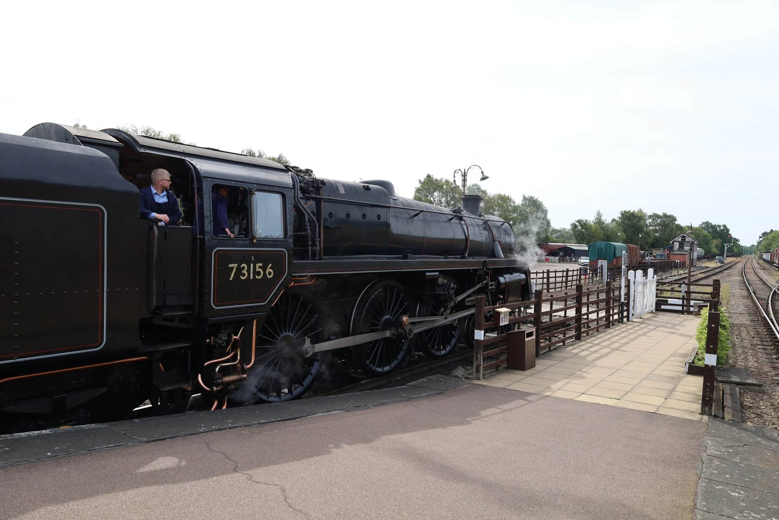 A vintage black steam locomotive number 73156 at a railway station, with passengers seen inside and steam rising from the engine, alongside railway tracks and station structures in the background.