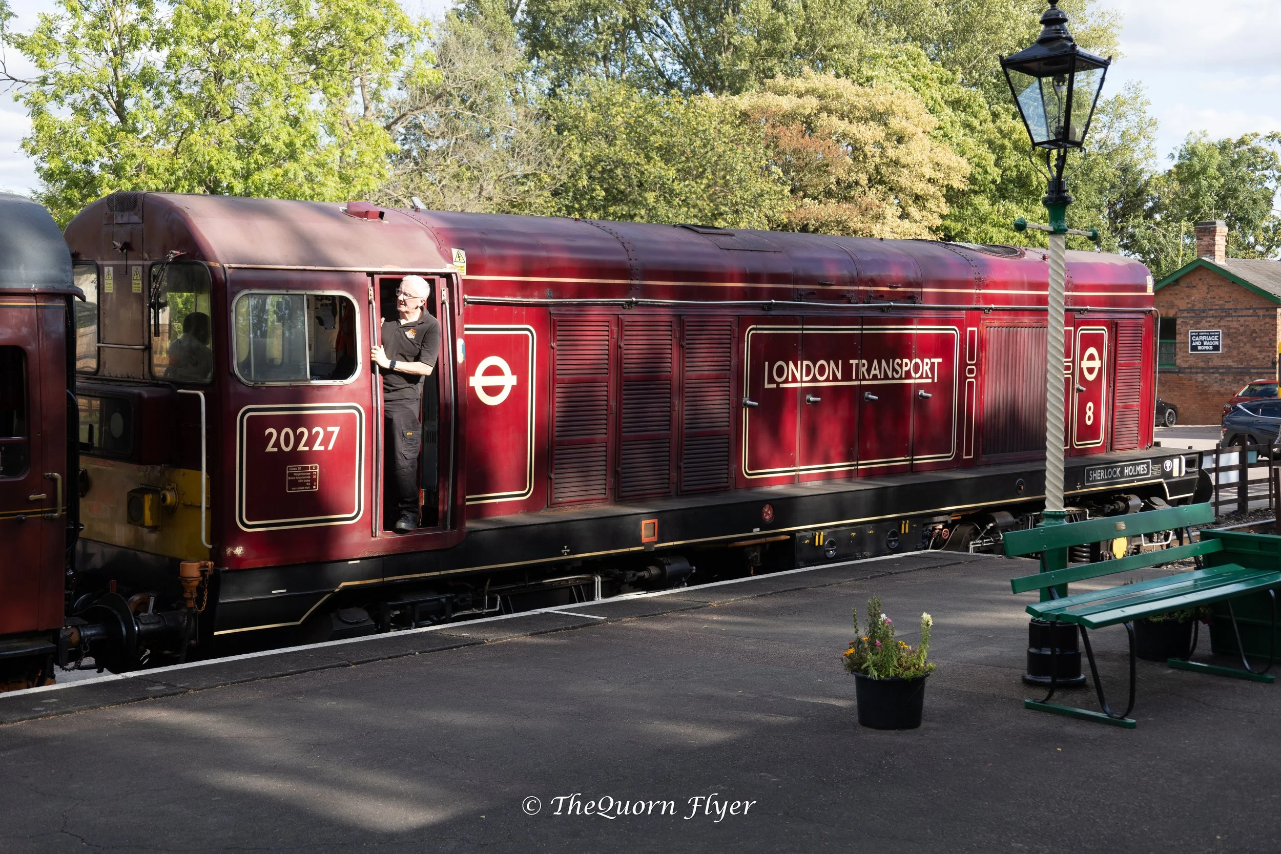 A vintage red London Transport train with a man standing in the driver's cabin, next to a black lamppost, some benches, and a flowerpot in a park-like setting with trees and buildings in the background.