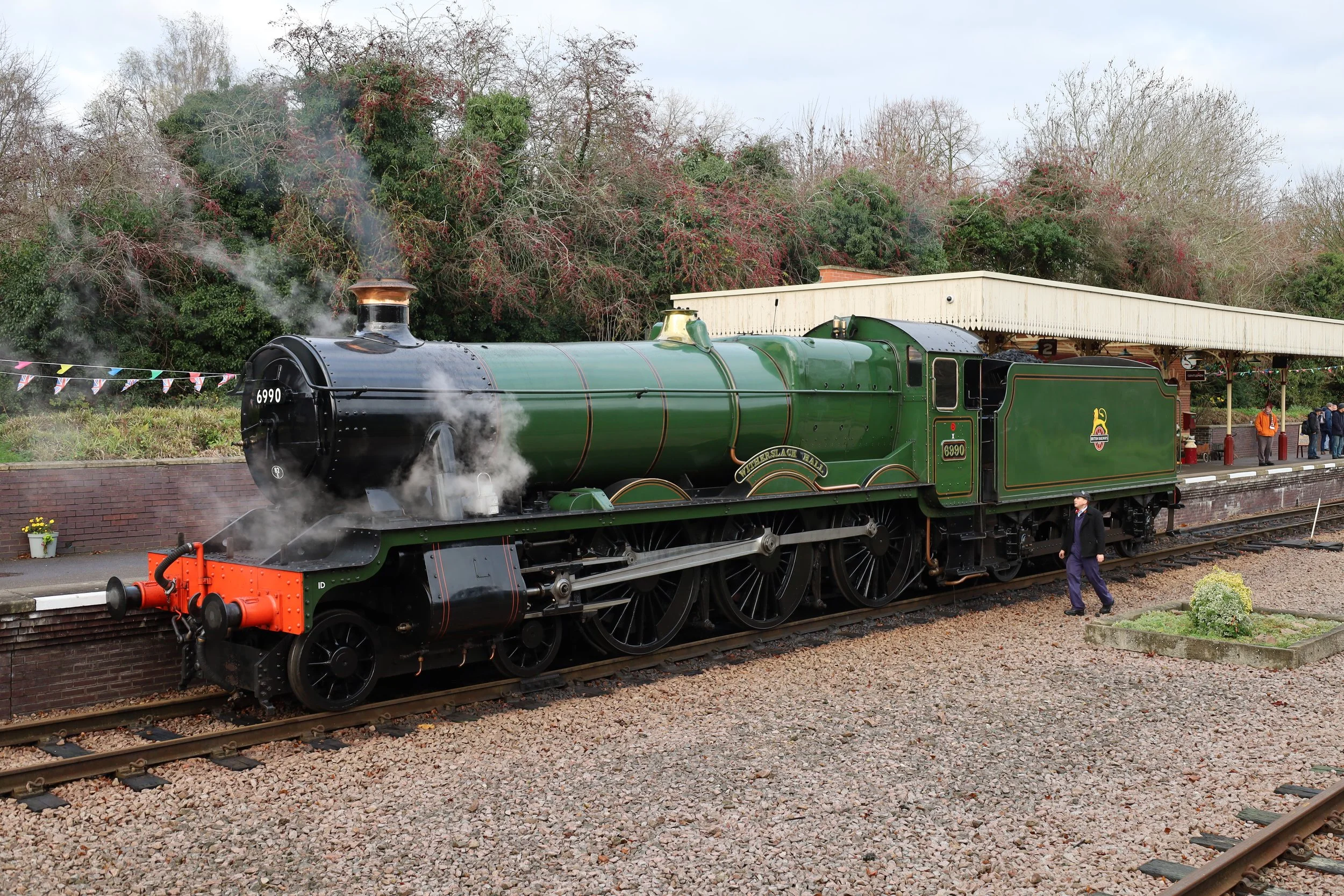 A vintage green steam locomotive with the number 6990 on the front and side, emitting steam, at a train station platform with people waiting, a canopy overhead, and trees in the background.