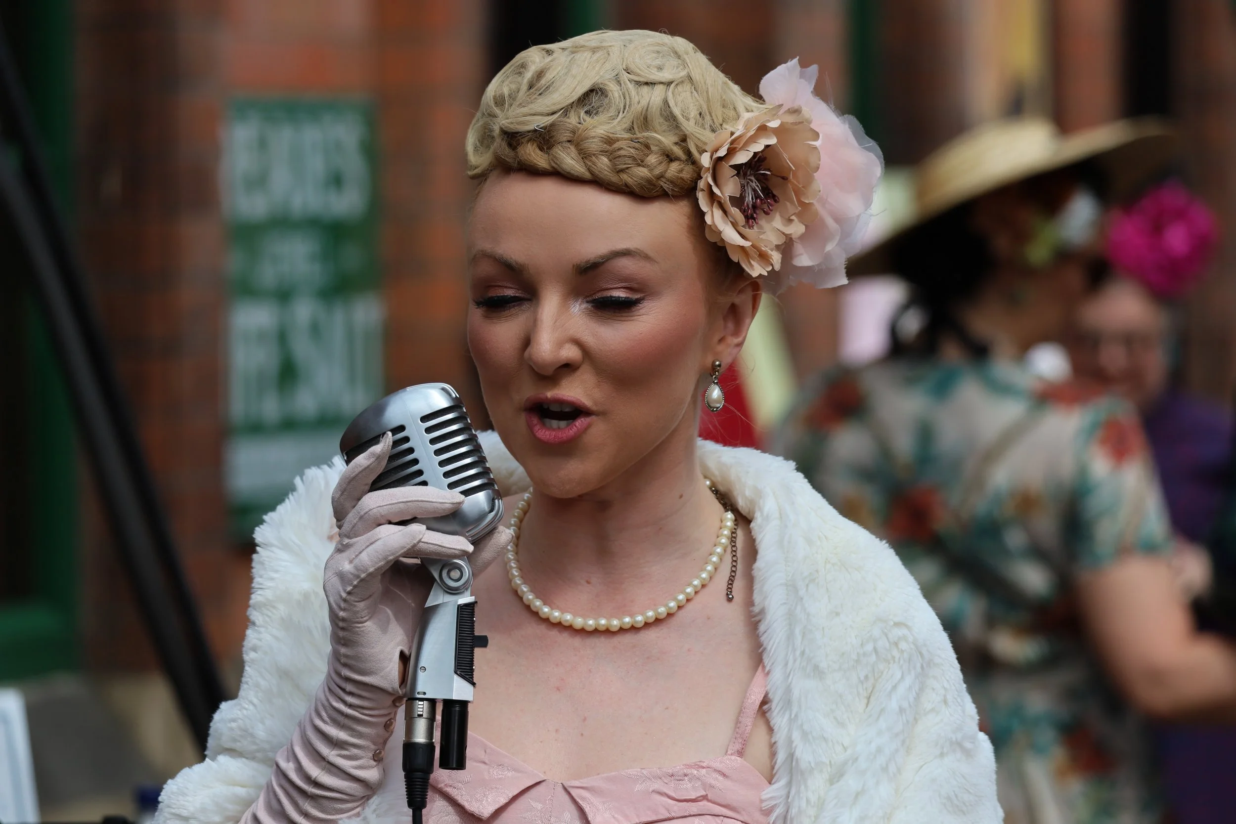 Woman with vintage hairstyle and pearl earrings singing into a microphone at an outdoor event, wearing a white fur coat and pink dress.