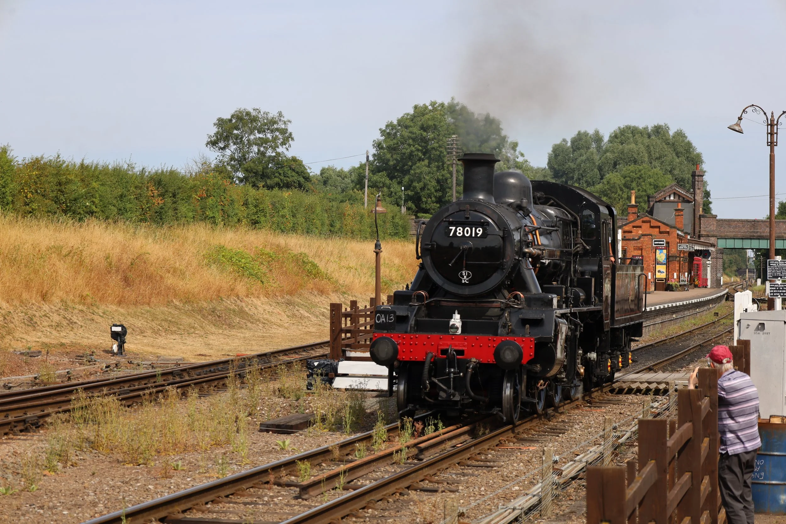 A vintage black steam locomotive train with the number 78019 on its front is on the train tracks at a station platform. There are people standing near the platform and a person taking a photo. The station has brick buildings, street lamps, and green trees in the background, with smoke coming out of the train's chimney.