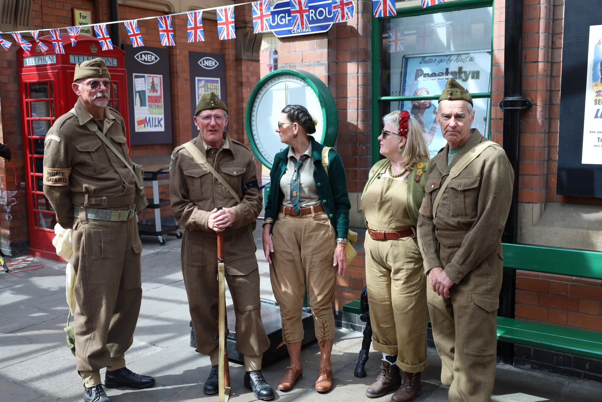 A group of five adults dressed in World War II-era military uniforms, standing on a city street decorated with Union Jack flags and bunting, with a red British telephone booth and a vintage clock behind them.