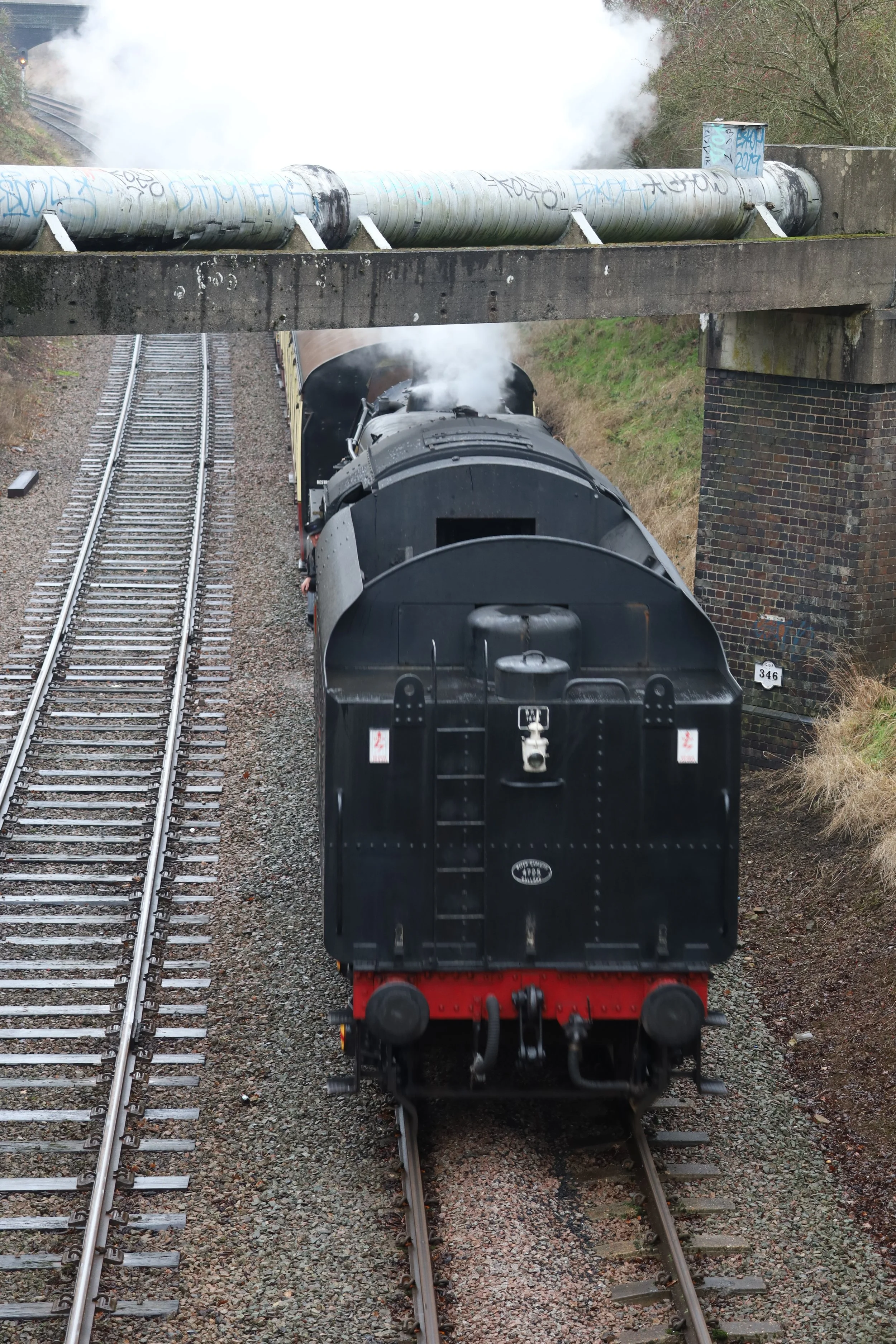 A black steam locomotive train traveling on railway tracks beneath an overpass with a large pipe emitting steam or smoke, with some graffiti on the overpass.