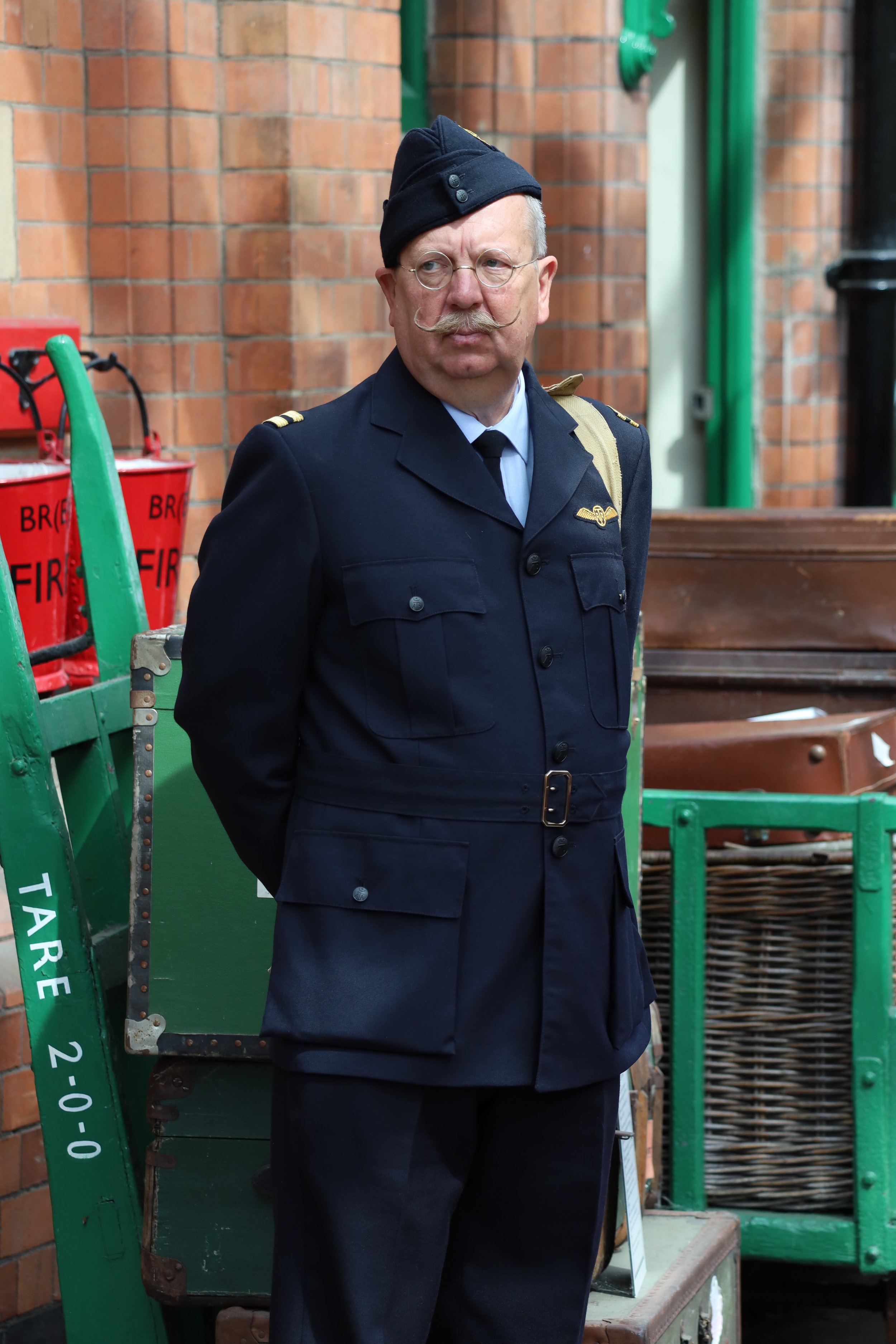 A man dressed as a pilot wearing a vintage military style uniform with a hat, glasses, and a handlebar mustache, standing outdoors against a brick wall with a green cart and other objects.