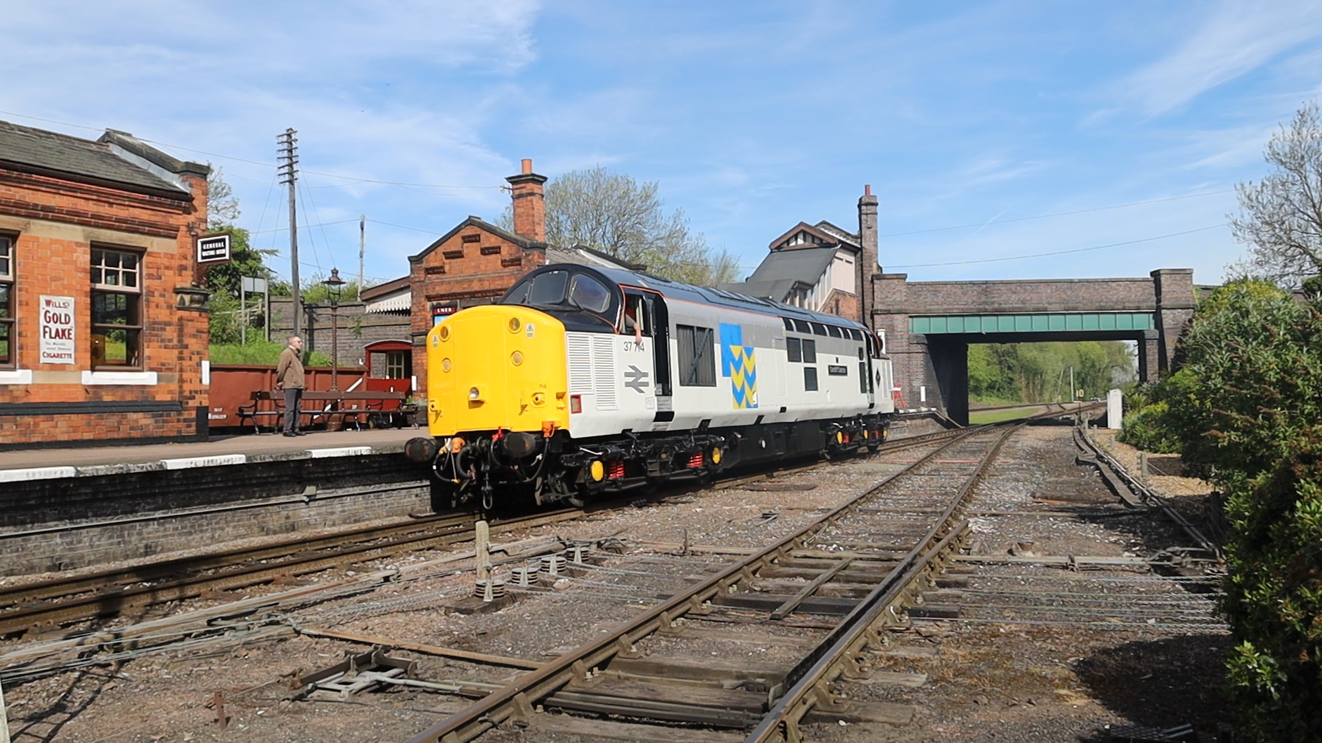 A train station with a vintage train on the tracks and a small platform, with a man waiting near a bench and brick buildings in the background under a blue sky.
