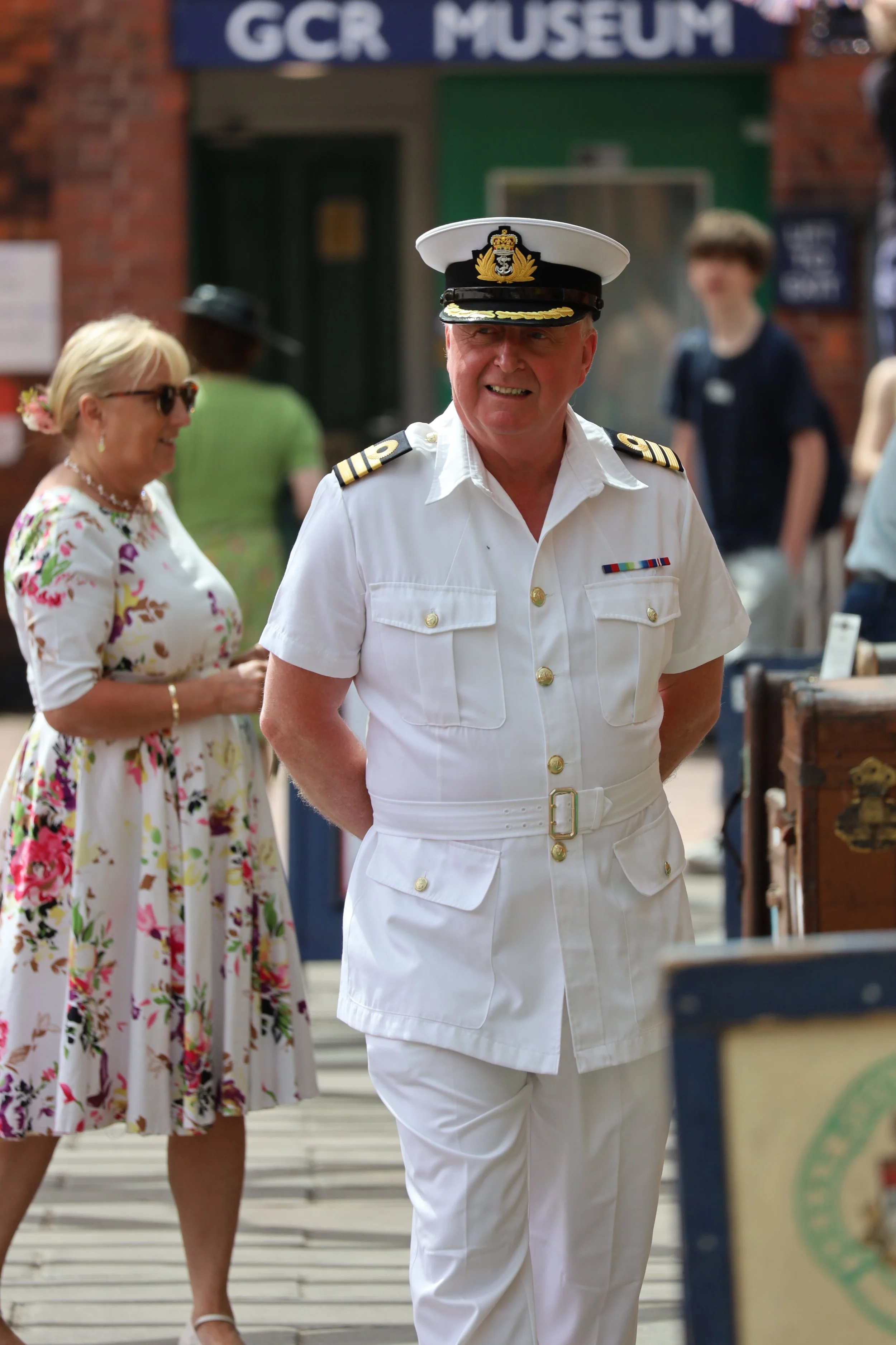 A man in a white naval uniform with a cap featuring a gold emblem, smiling as he walks outdoors, with a woman in a floral dress and sunglasses behind him, and other people in casual clothing around, near a brick building and a sign that reads GCR MUSEUM.