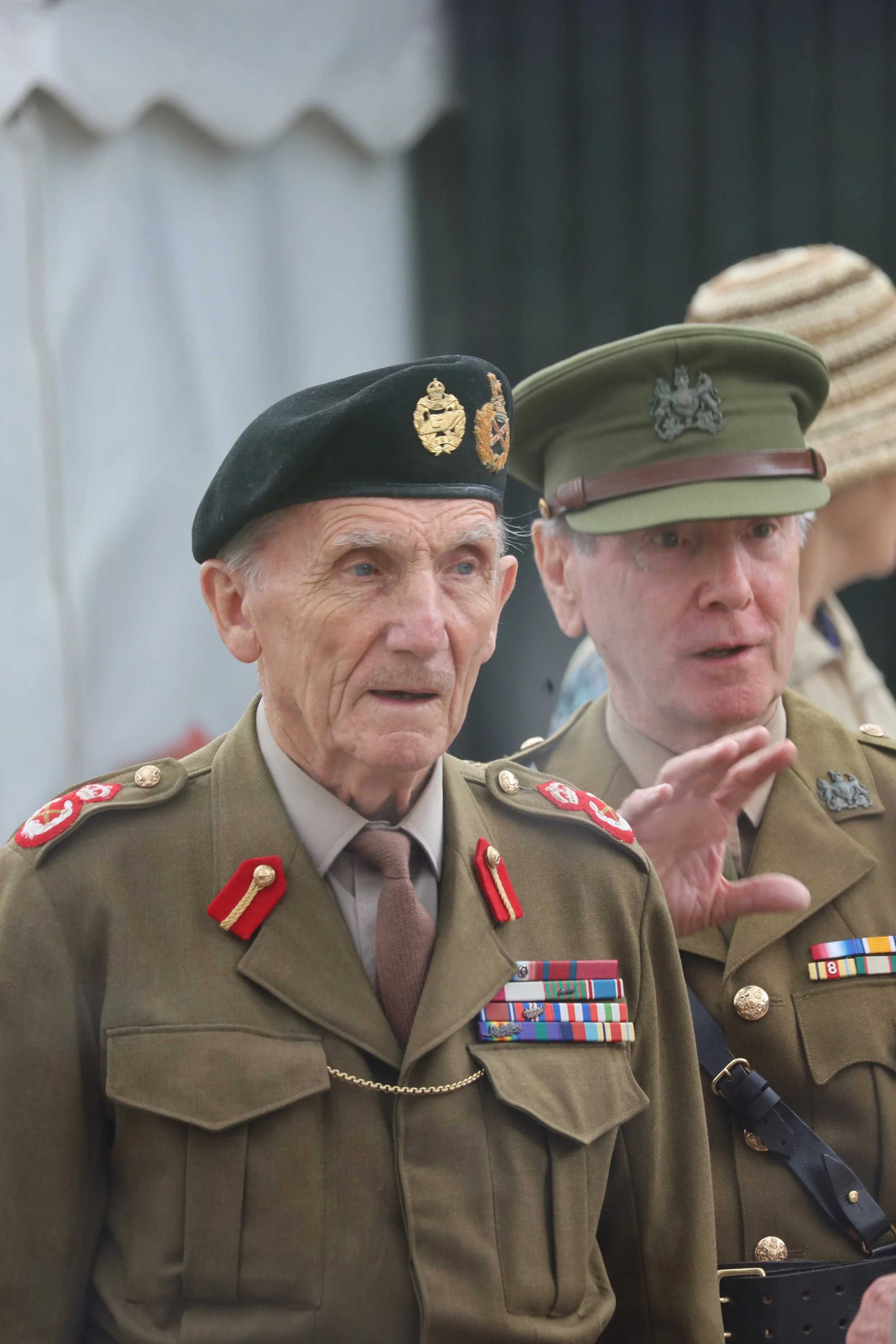 Two older men in military uniforms are engaged in a conversation. One wears a black beret with military insignia, the other a green cap with a crest. They are decorated with various medals and ribbons.
