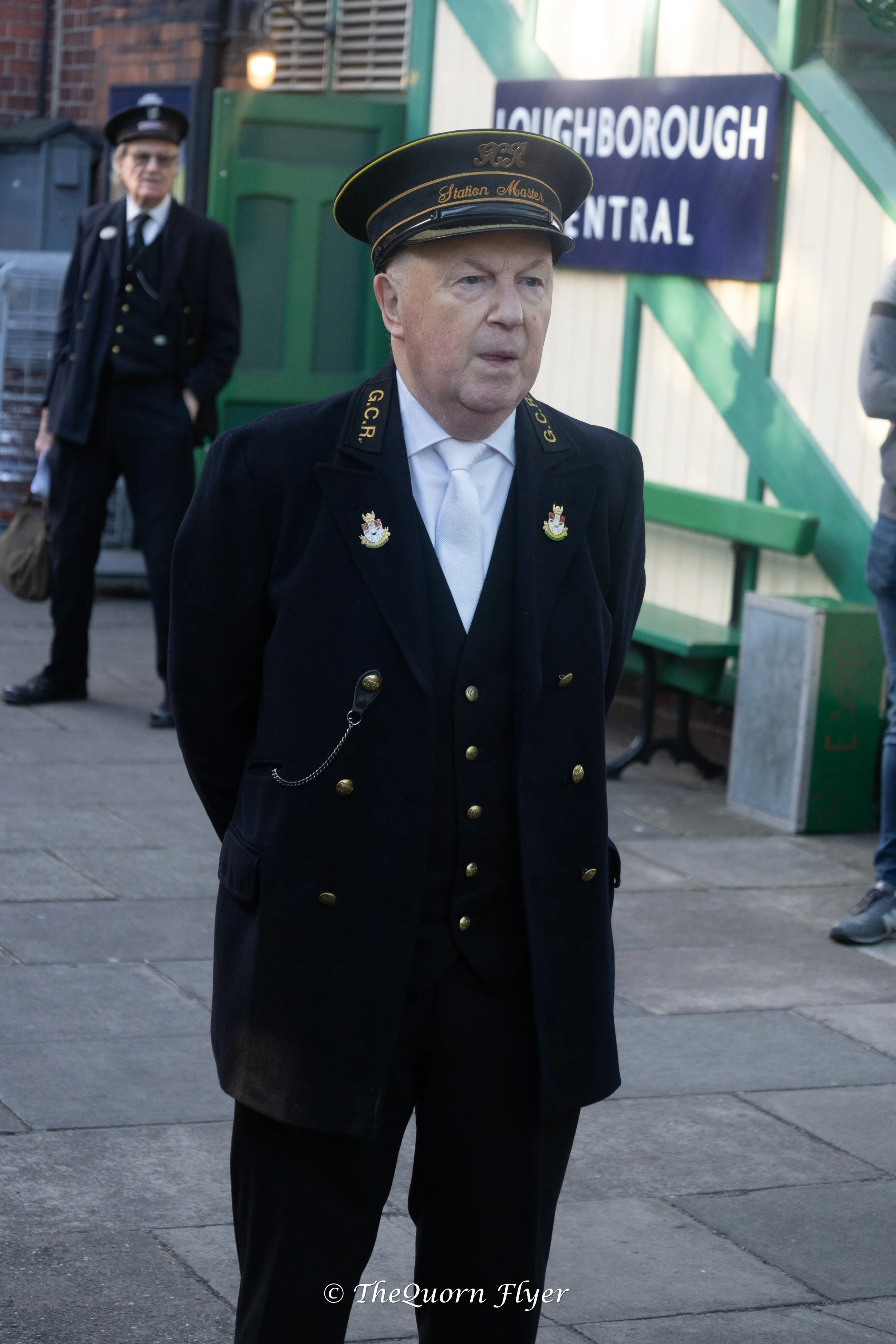 A man dressed as a train conductor standing outdoors in front of a building with a blue sign that reads 'Loughborough Central'. He is wearing a black uniform and cap with gold details, and appears to be waiting or observing something. Behind him is another person dressed in a dark suit and hat, standing with hands in pockets. There is a green bench and a trash can nearby.