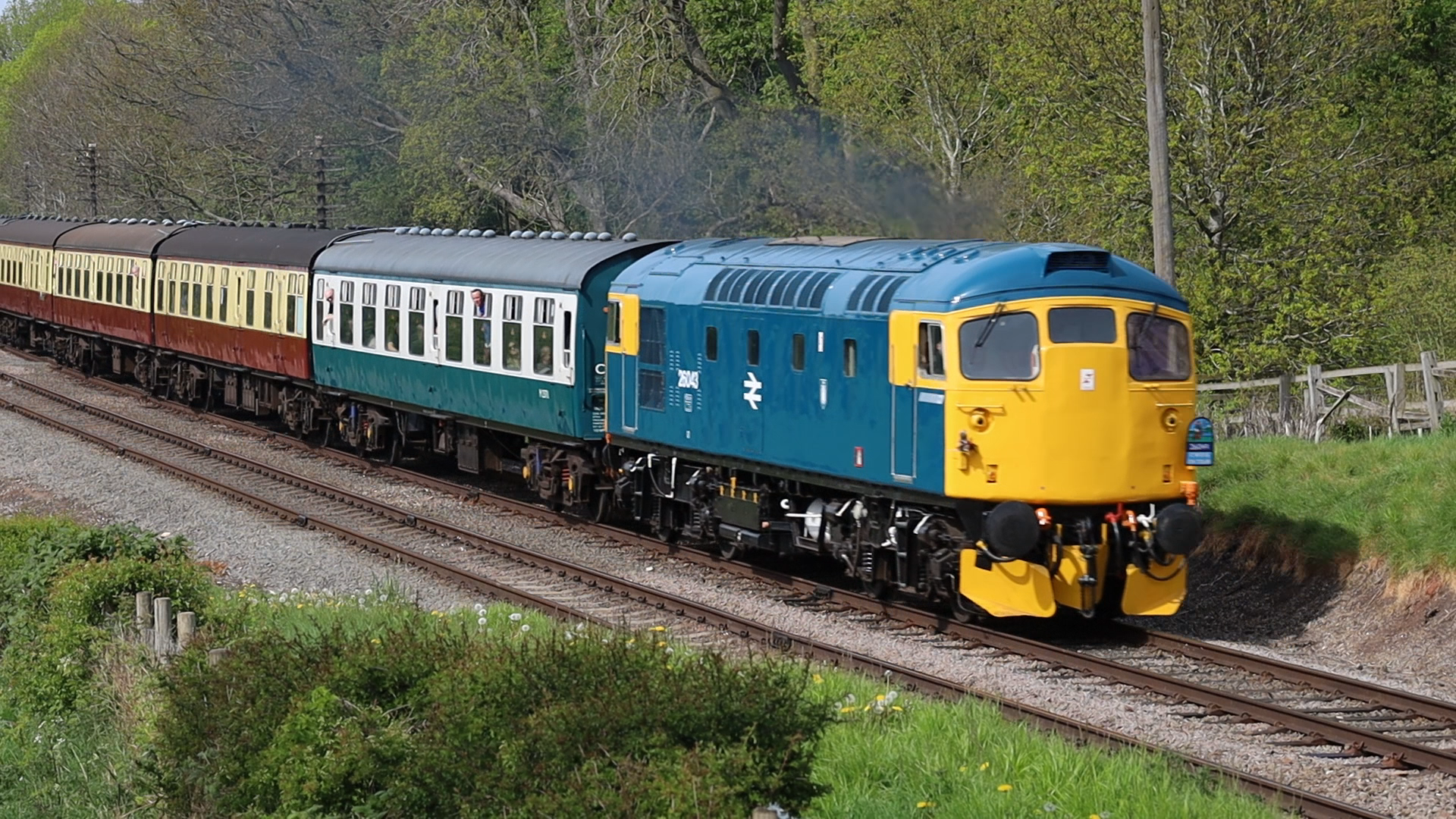 A vintage blue and yellow diesel locomotive pulling a passenger train through a rural area with greenery and trees.