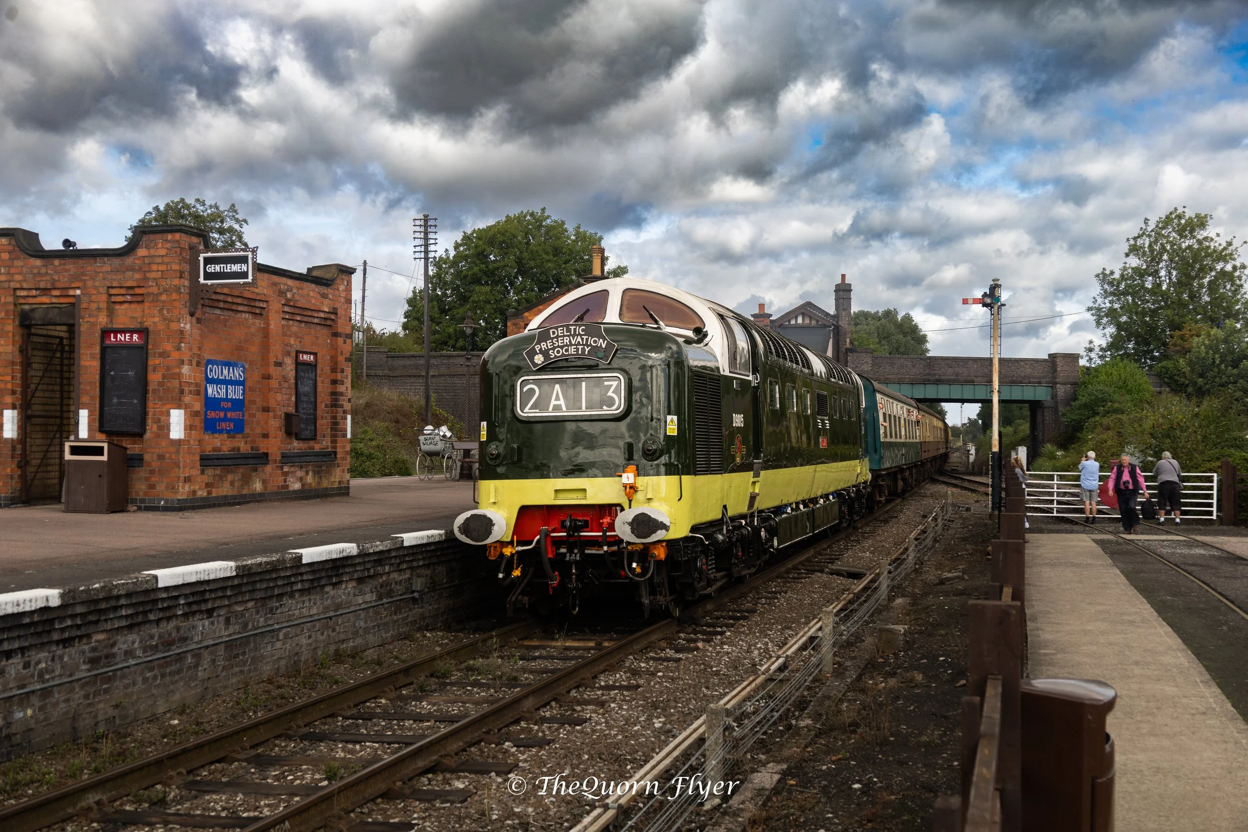 A vintage green and yellow train stopped at a small train station on a cloudy day. There is a brick building with a sign that says 'Gentlemen' and posters on the wall. Several people are walking on the platform, and trees and a bridge are in the background.