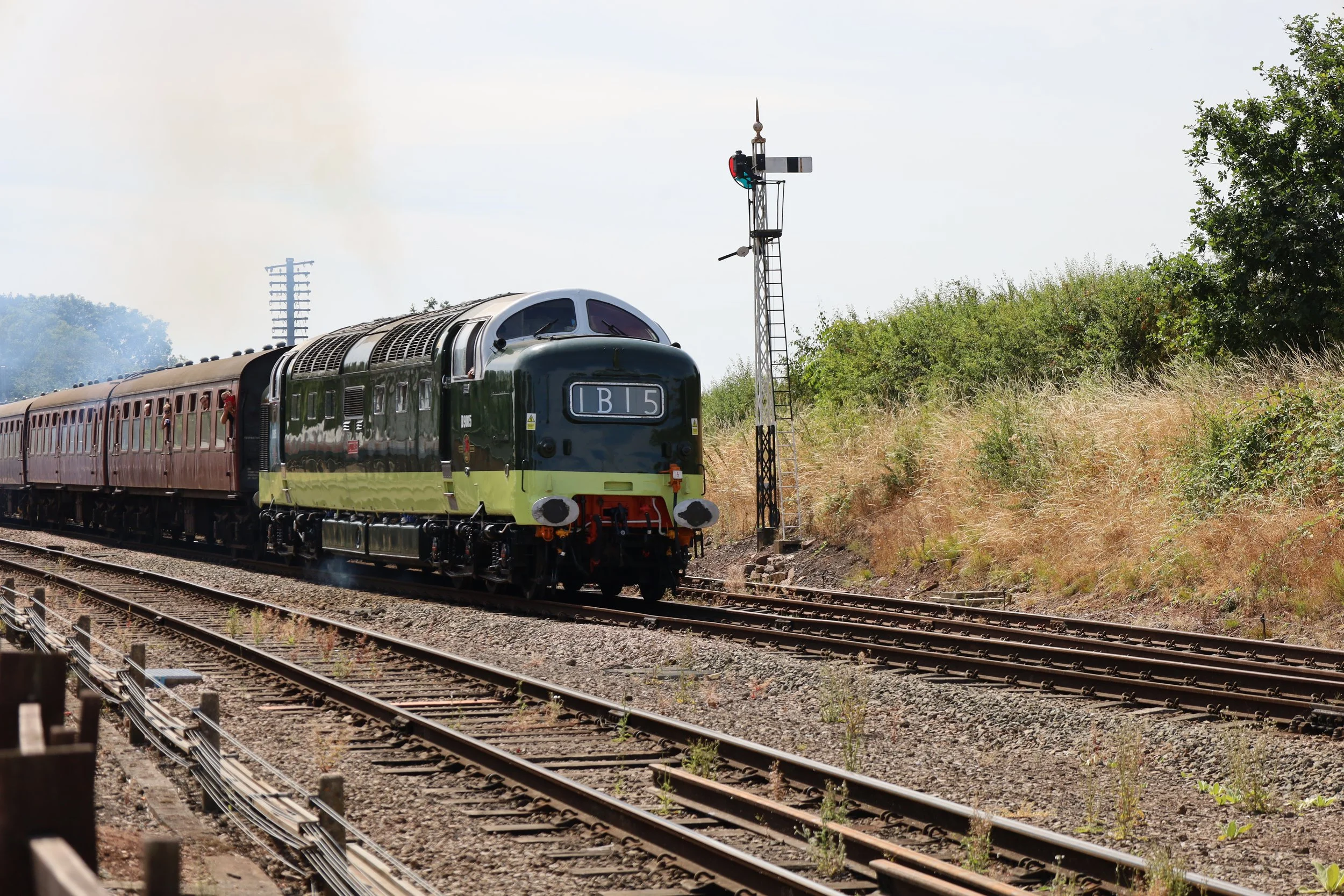A vintage green train traveling on railway tracks with a semaphore signal and greenery in the background.