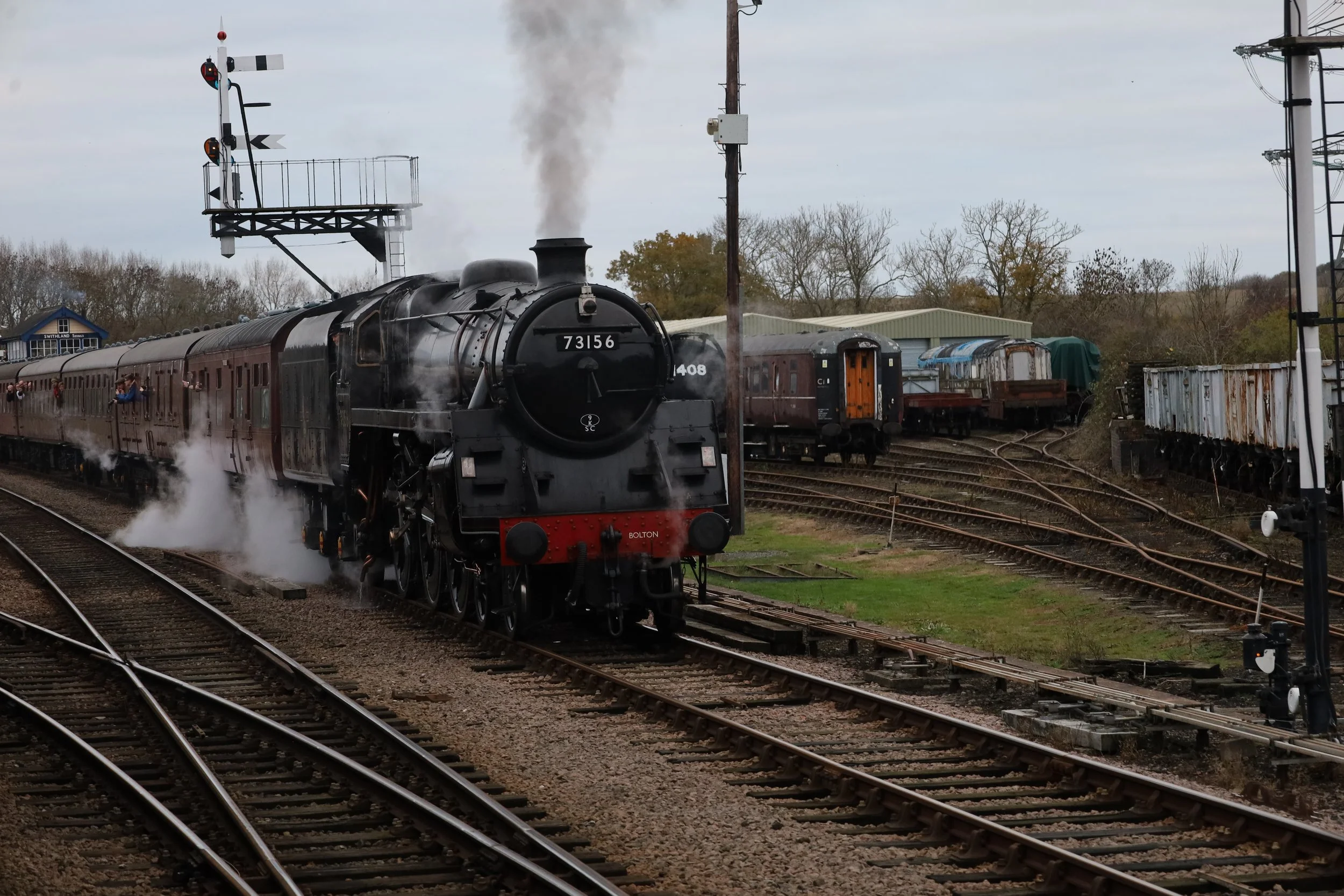 A vintage black steam locomotive with the number 73156 making smoke as it moves along train tracks, with several passenger cars attached. There are additional train cars and a signaling system with lights in the background, along with a small building and trees, under a cloudy sky.
