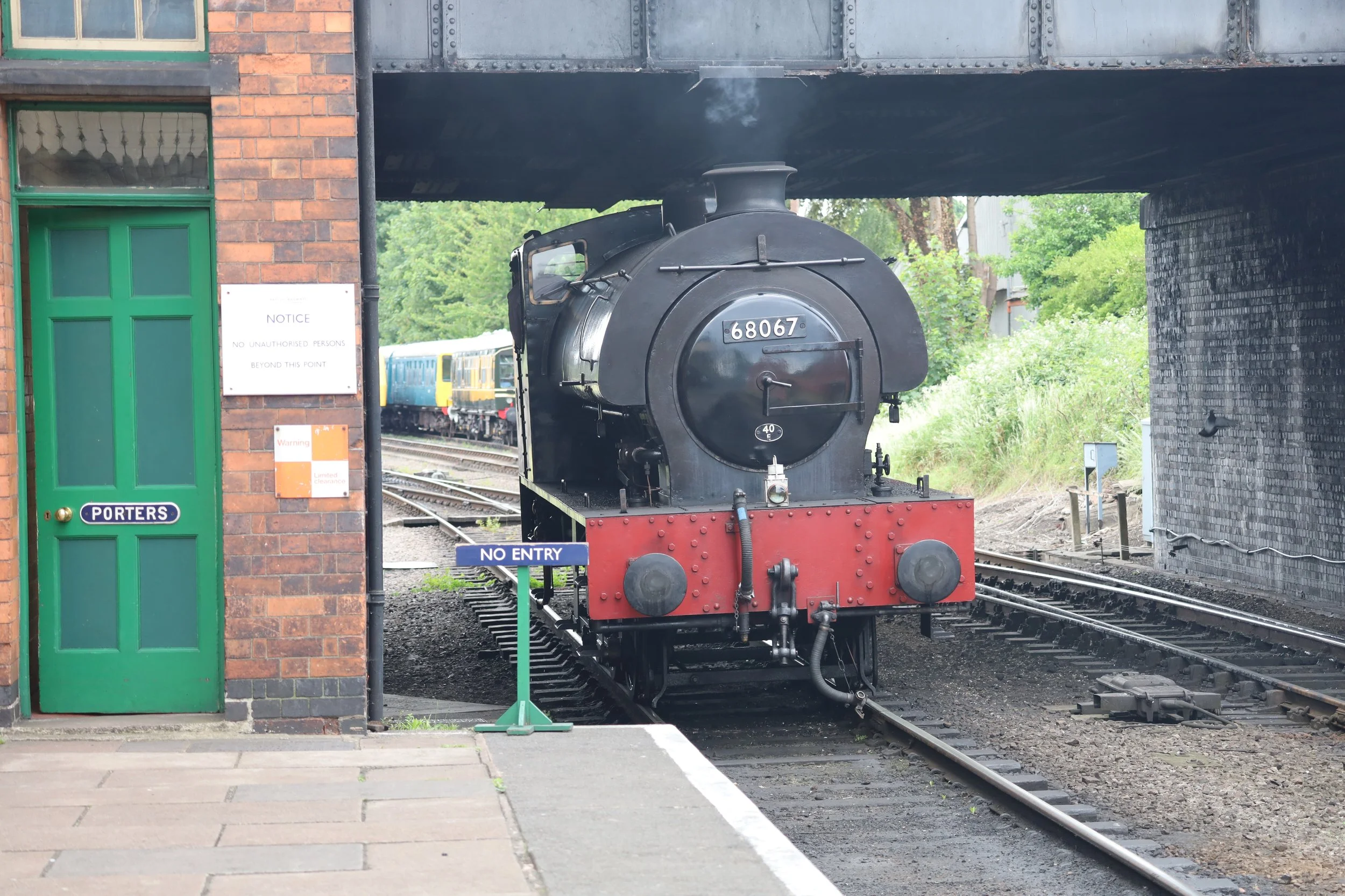 A vintage steam train with the number 68067 is exiting a brick railway station under a bridge. A sign near the train reads "No Entry" and another sign says "Porters." The train is passing on tracks surrounded by green trees.