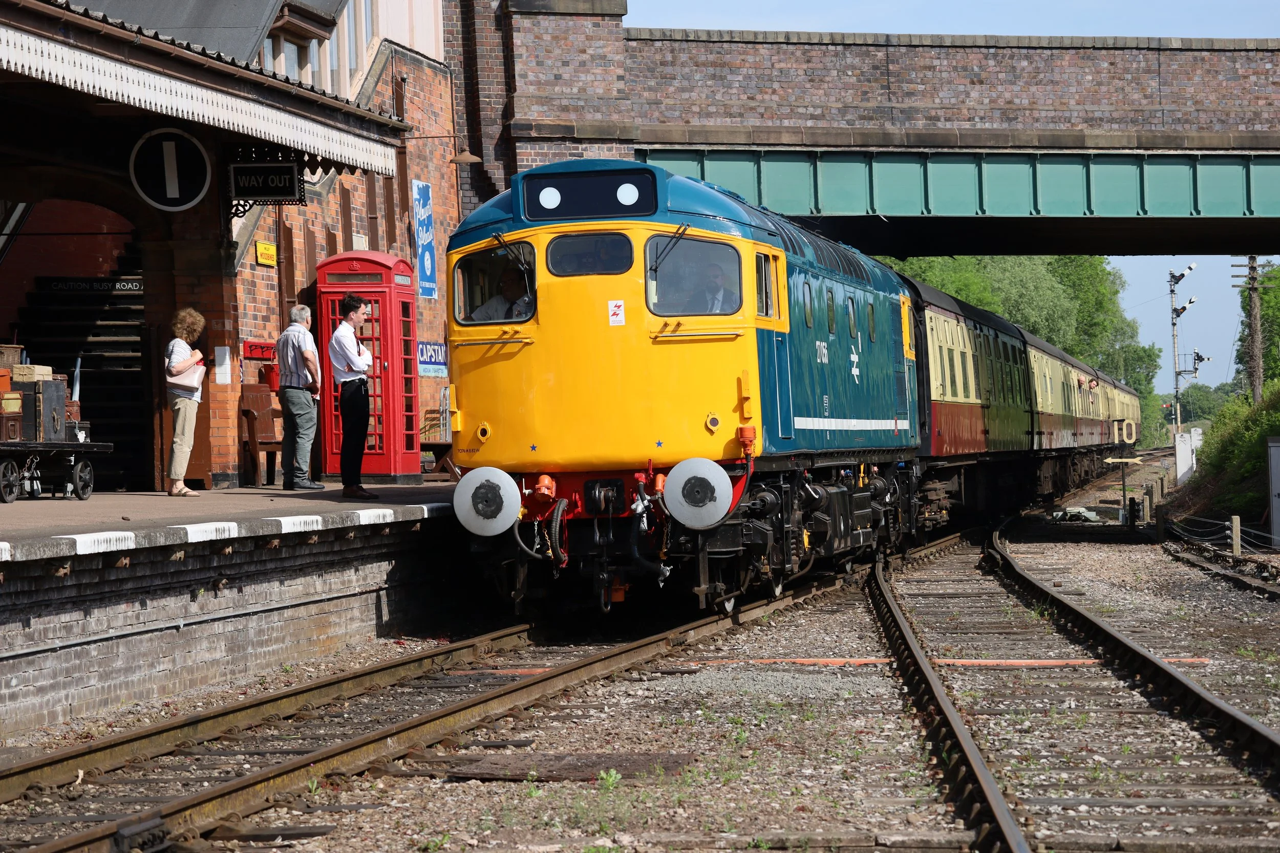 A colorful vintage train with blue and yellow front section near a brick train station platform. Several people are waiting near a red British telephone booth and signage. The scene is outdoors with track rails and semaphore signals in the background.