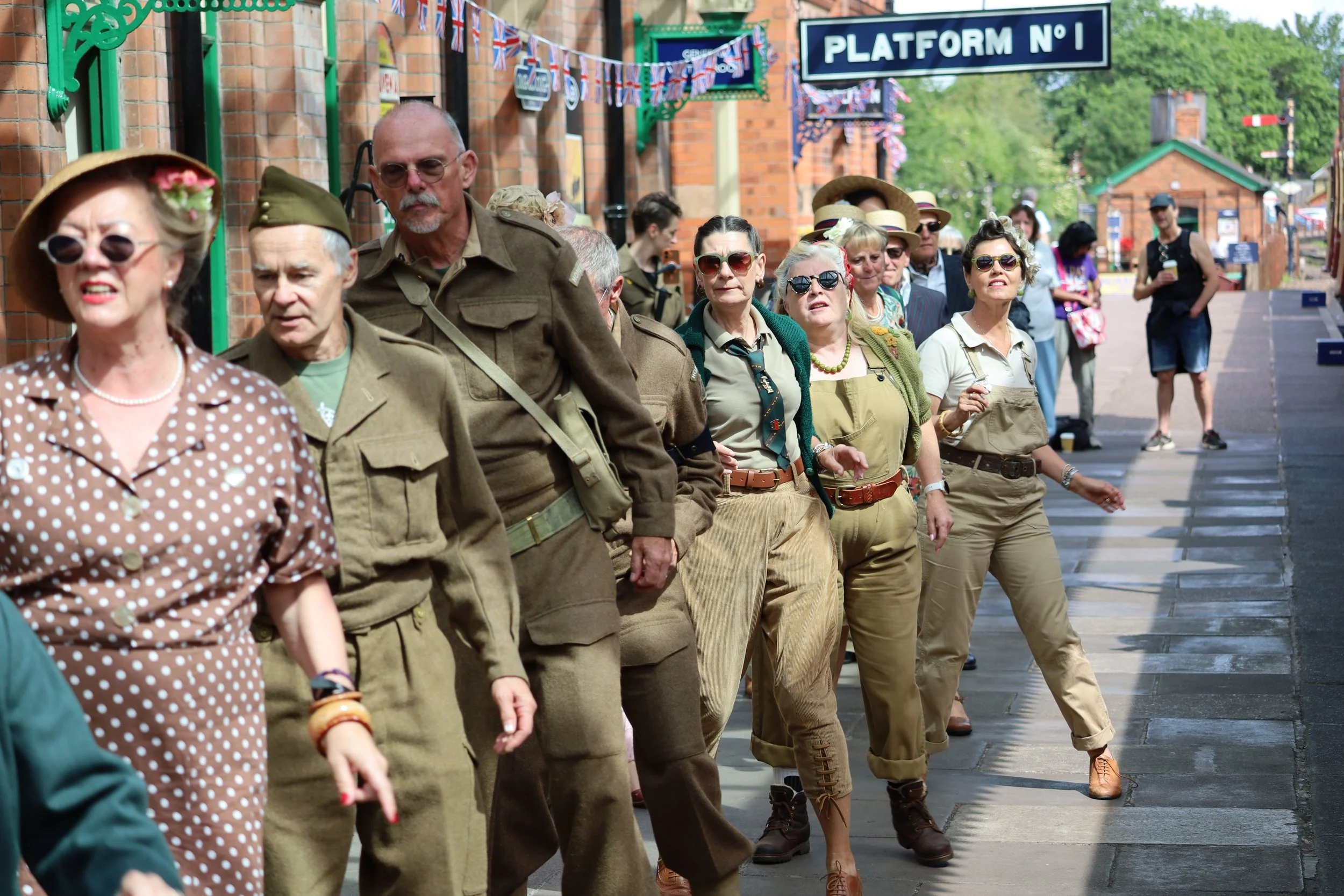 A group of people dressed in vintage military-style clothing and 1940s fashion, standing in line outside a building with a sign reading 'Platform No. 1,' decorated with British flags and bunting, suggesting a nostalgic or historical event.