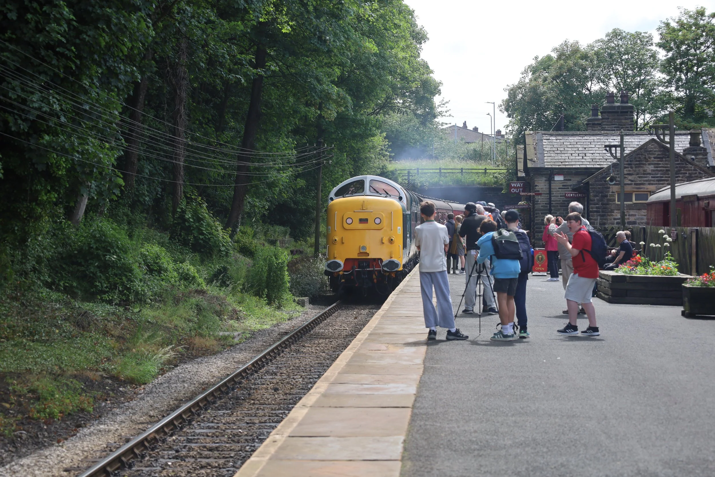 A vintage train arrives at a small station platform filled with people, with trees and a stone building in the background.