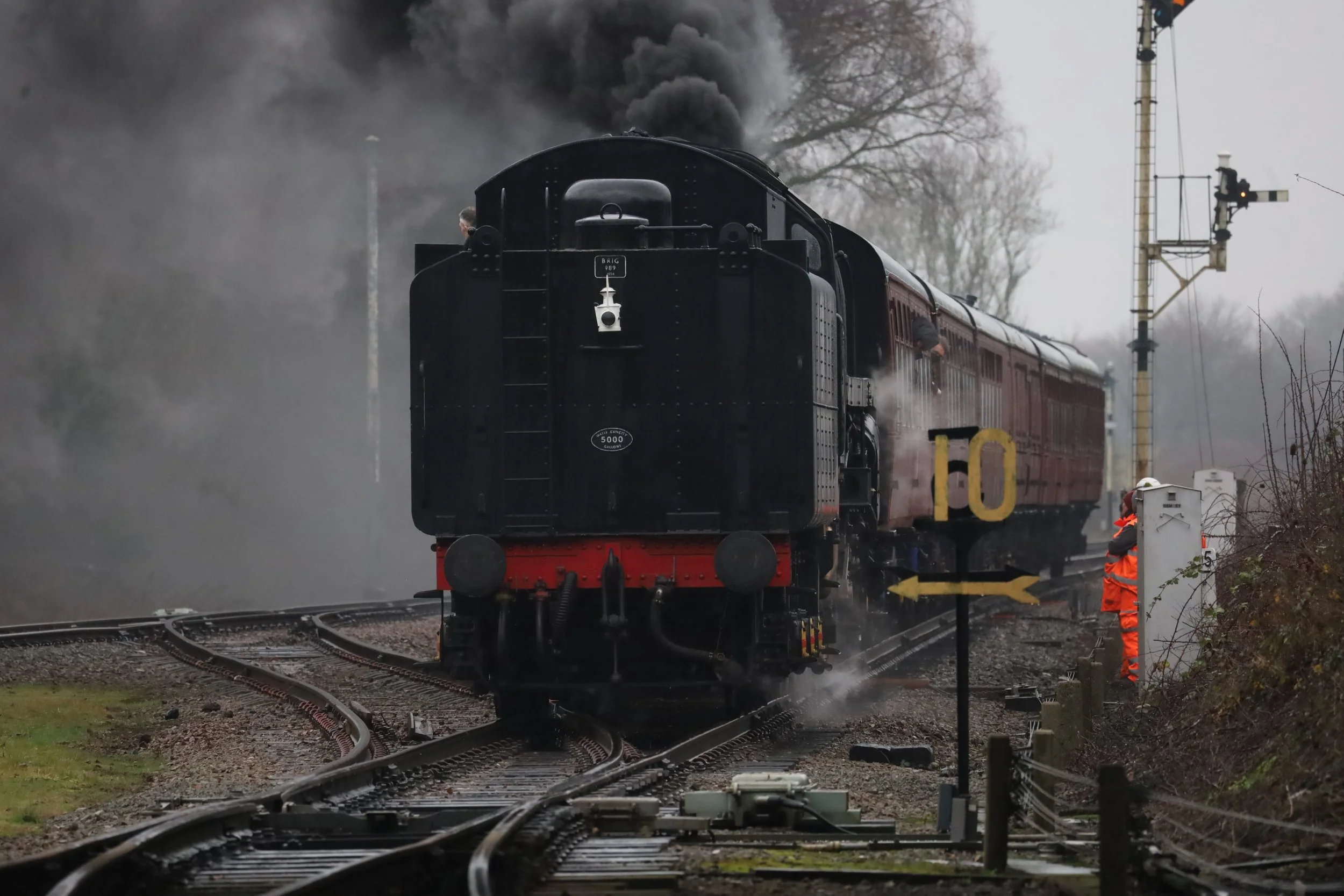 A vintage steam locomotive with black and red coloring pulling several passenger cars on railroad tracks. Smoke is billowing from the engine's chimney, and train workers in safety vests are working near the track. There is a railway signal and switch equipment nearby, with a gray, overcast sky in the background.