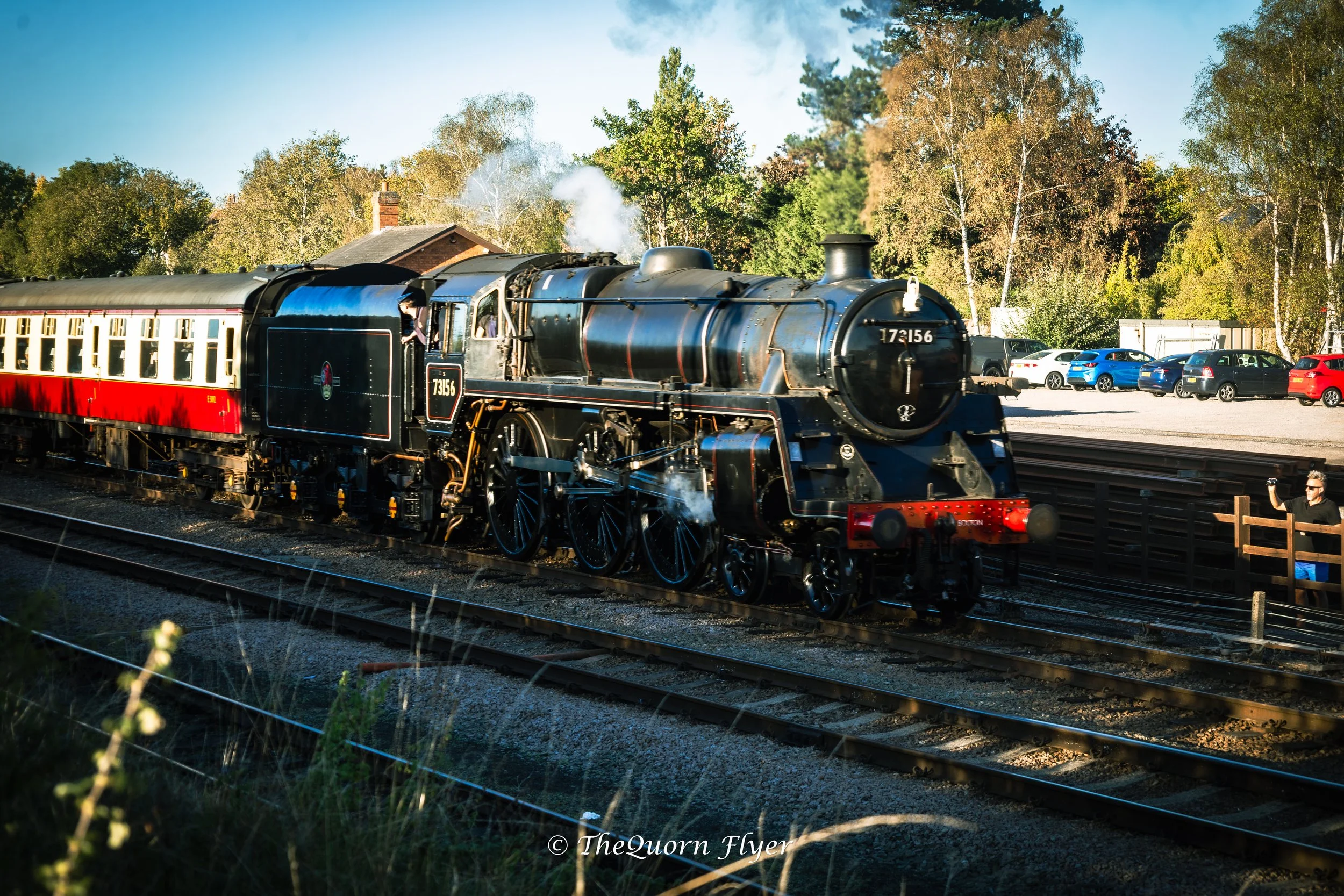 A vintage black steam locomotive train with the number 72156 on its front, pulling passenger cars along a railway track on a clear day. There are some cars parked on the side and a person visible near the train, with a background of trees and a small building.