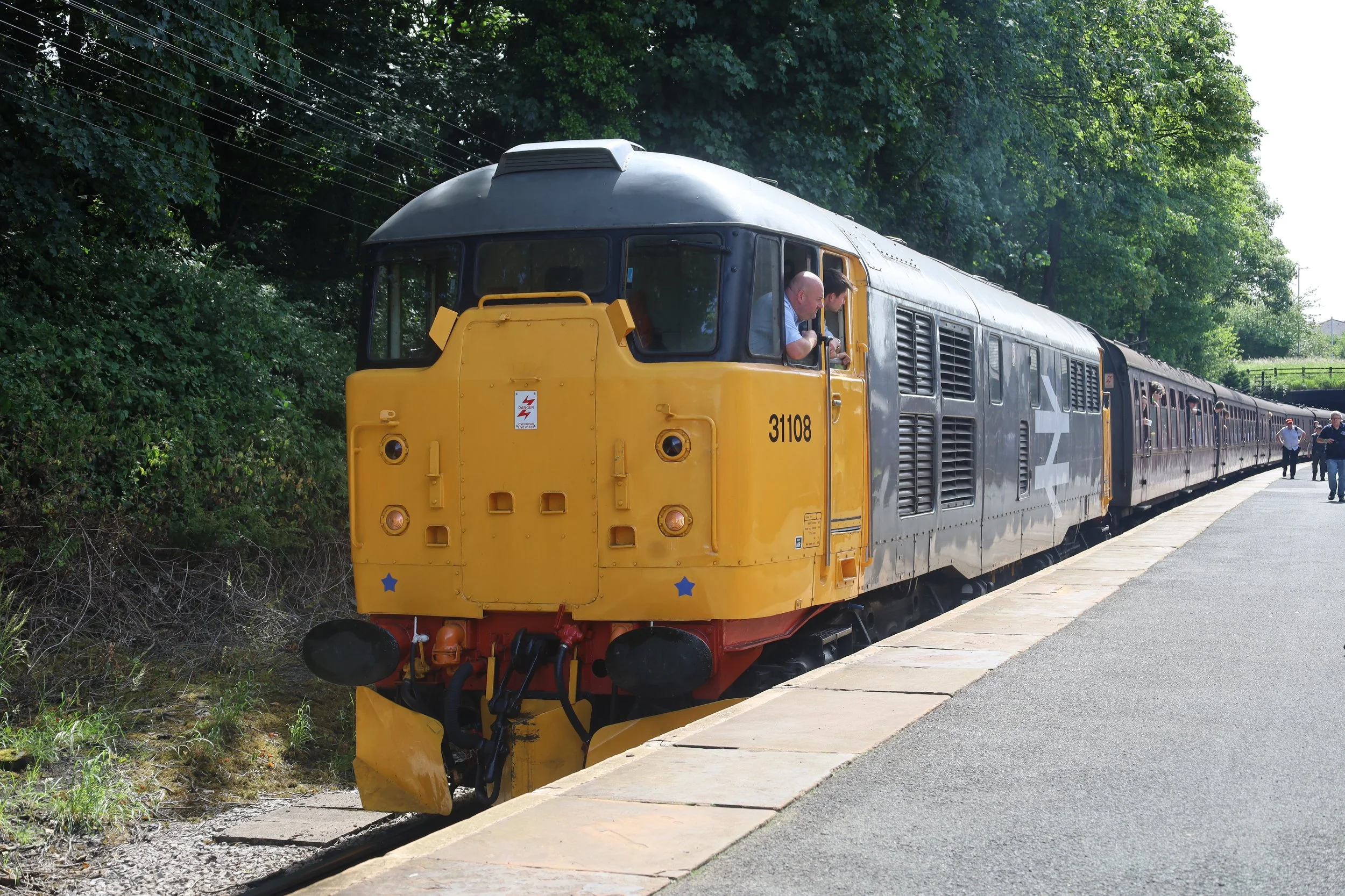 A yellow train engine with the number 31108 is on a train platform next to a grassy area with trees. There are train cars attached behind it, and a few people are walking on the platform.