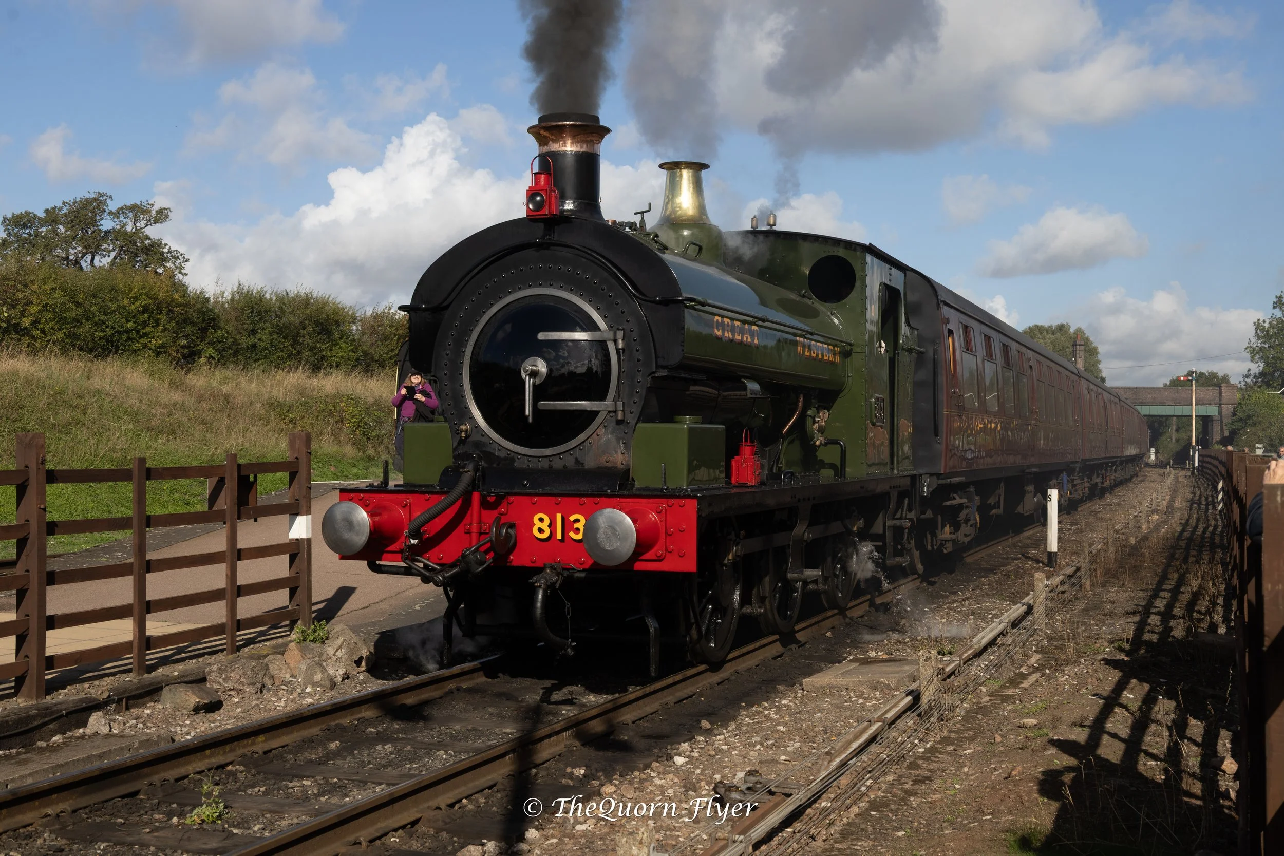 A vintage steam locomotive pulling train cars on railway tracks, with a person standing nearby taking a photo. The locomotive is black and green with the number 813 on the front, emitting black smoke, under a partly cloudy sky.