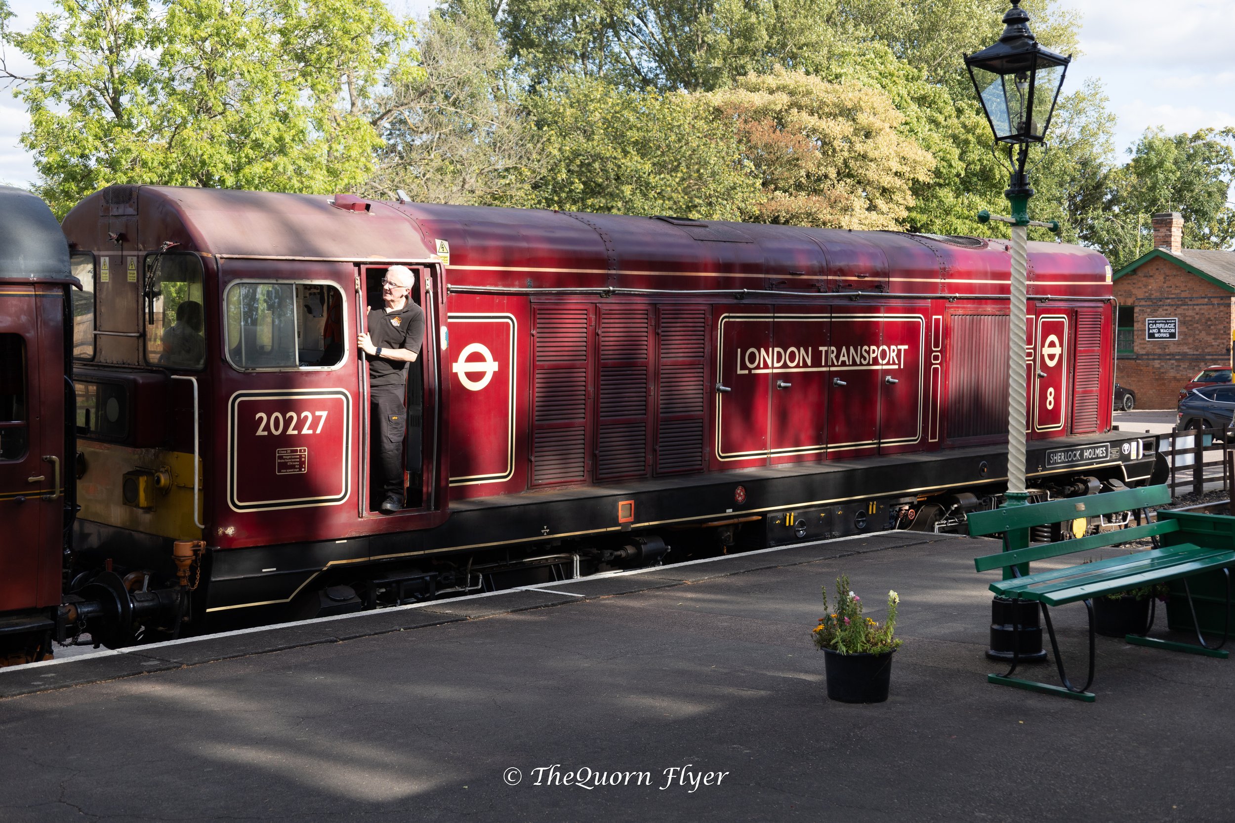 A vintage red train with the words 'London Transport' on its side, parked on a track, with a man standing at the front, surrounded by green trees and a building in the background.