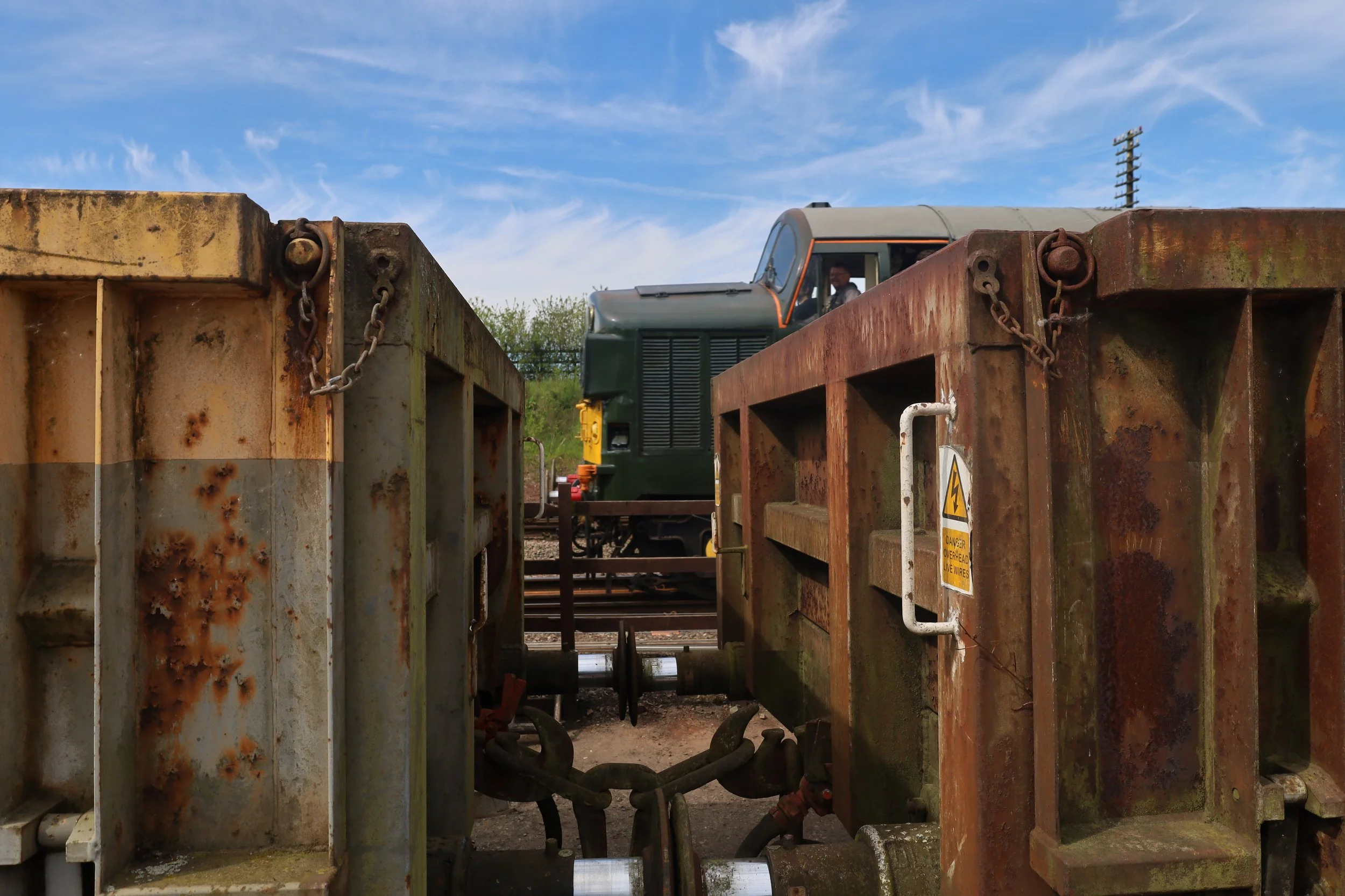 Rusty metal containers with chains, a train, and a conductor visible through a window, set against a blue sky with scattered clouds.