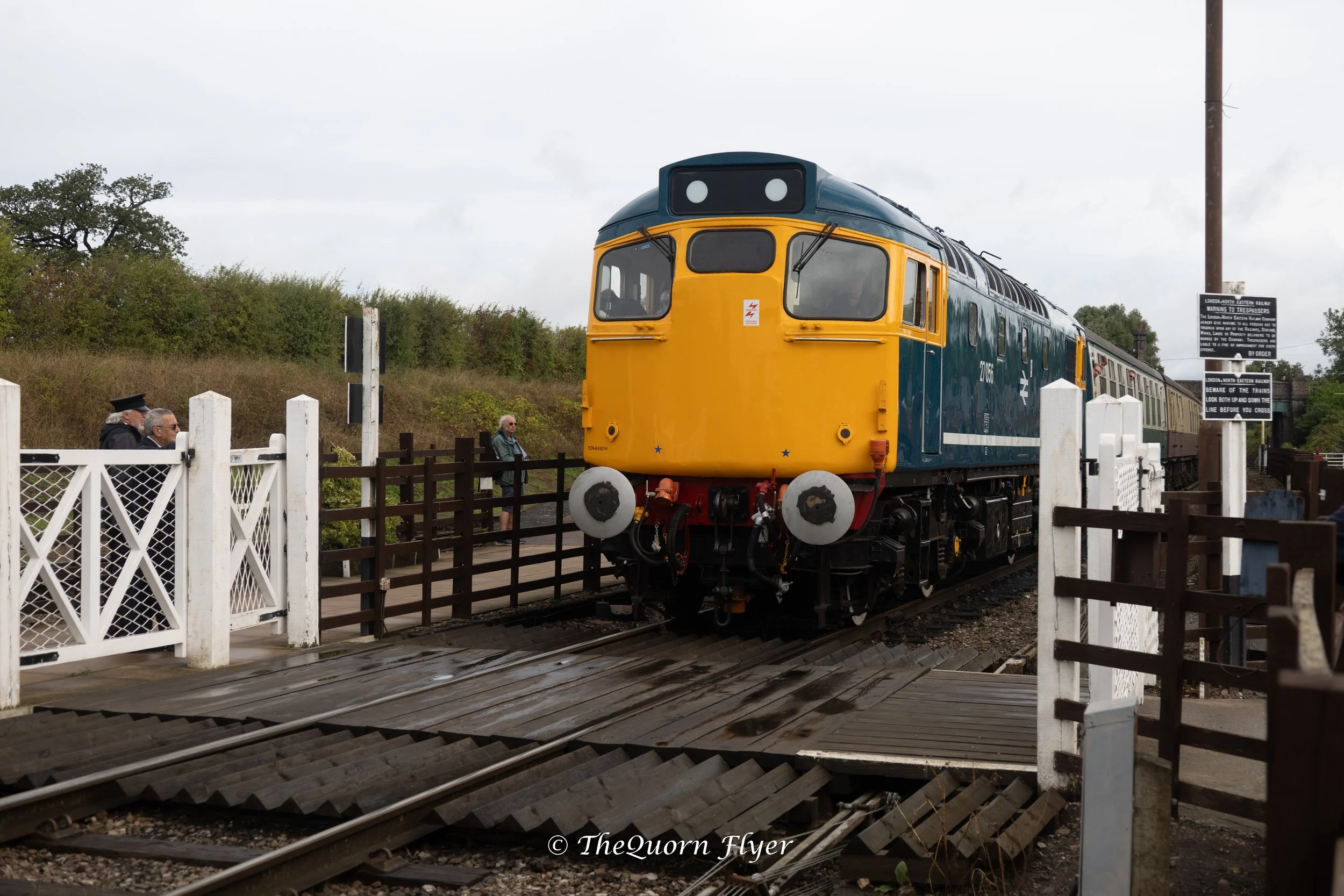 A vintage train painted in blue and yellow is passing by a gated crossing with a handful of pedestrians watching. There is a sign with safety warnings near the crossing.
