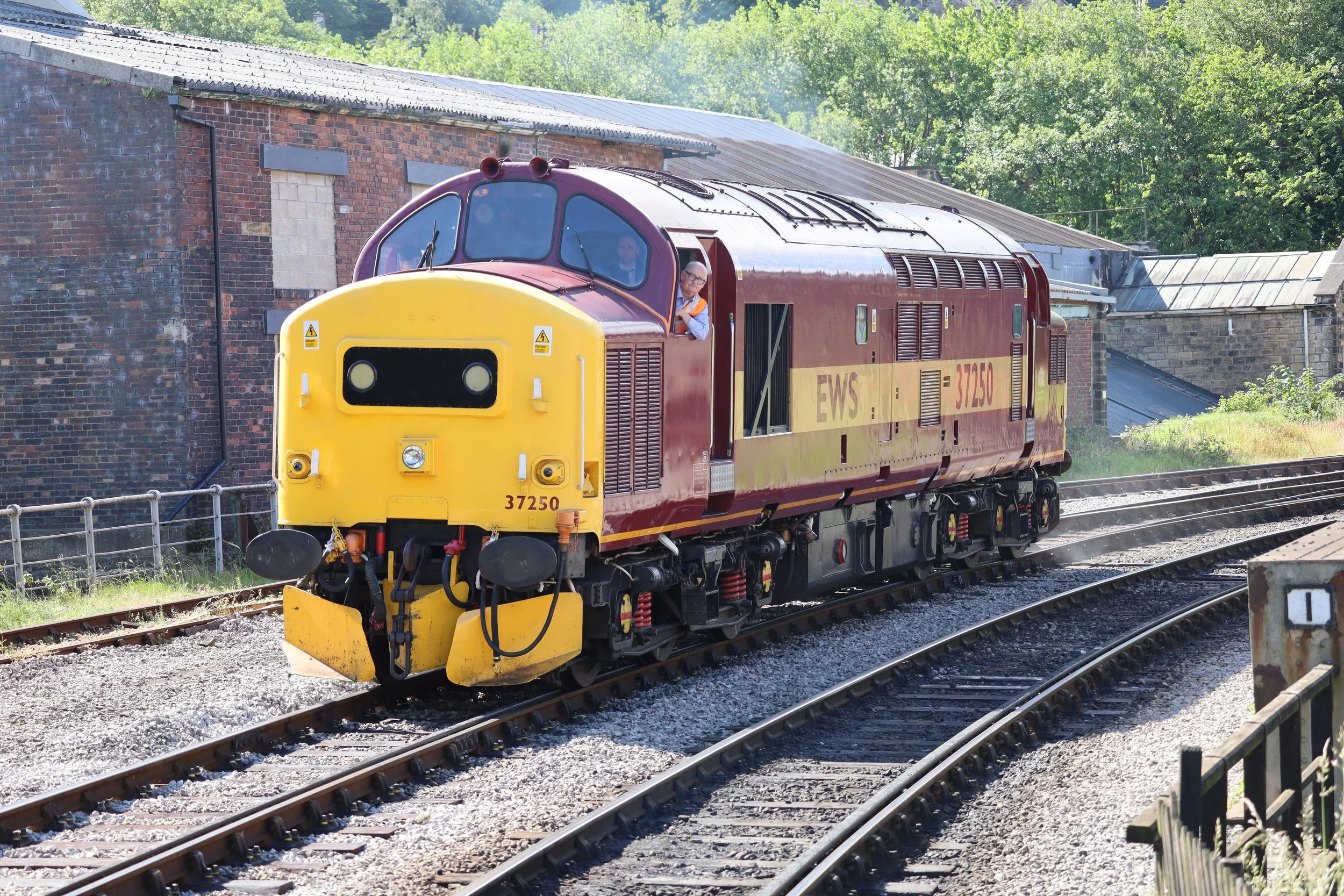 A red and yellow locomotive train moving on railway tracks past a brick building with a sloped roof, with lush green trees in the background.