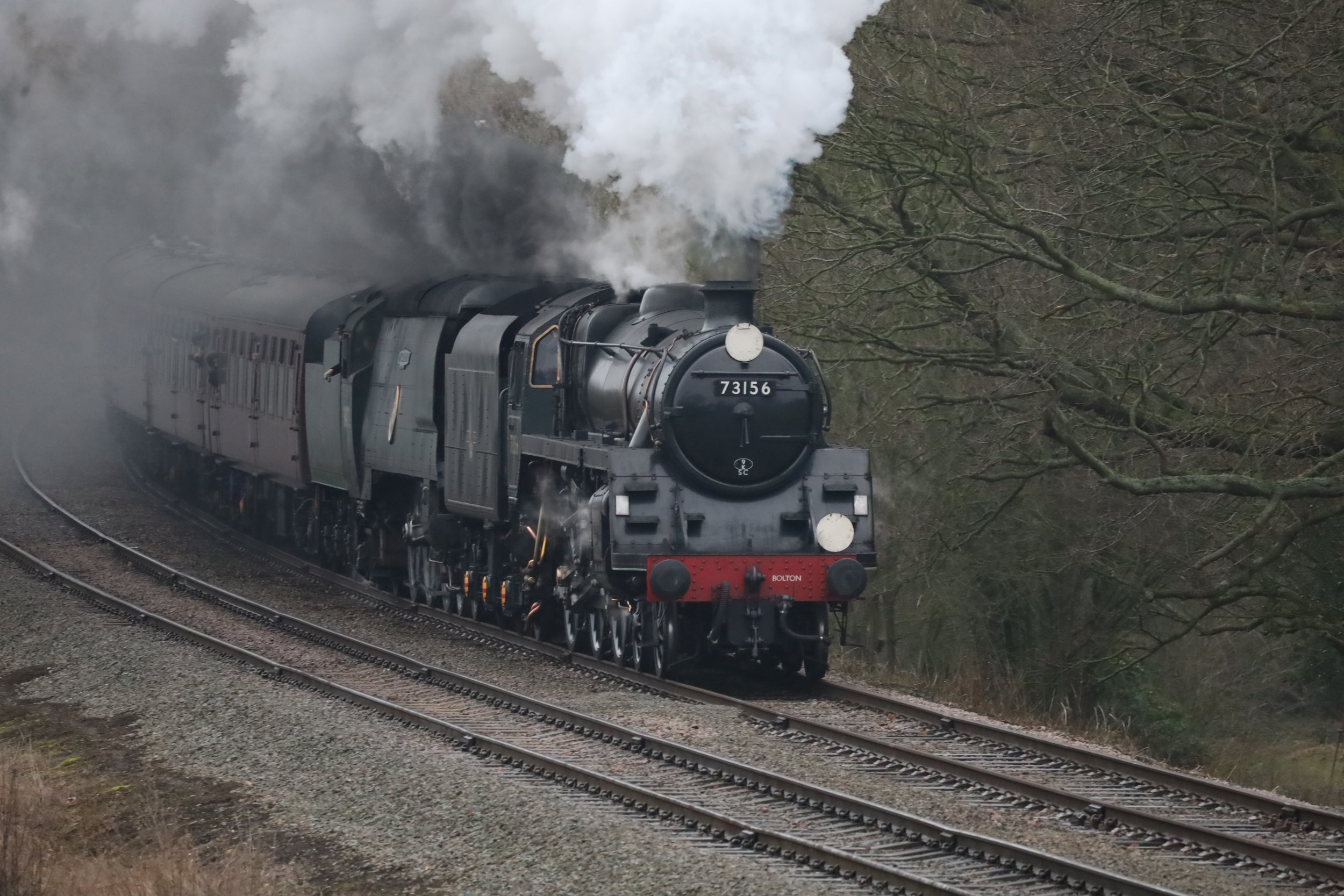 A vintage steam locomotive train traveling on tracks through a wooded area, emitting white smoke from its chimney.