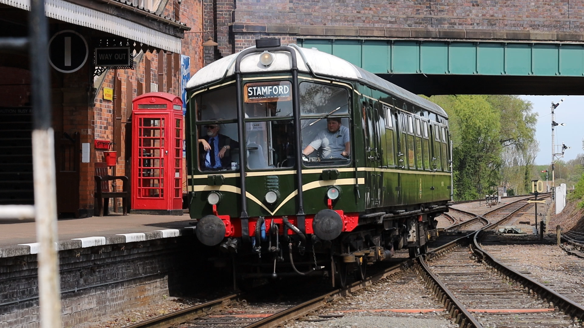 A vintage green and cream tram with a sign indicating it is headed to Stamford, arriving at a station platform. Behind the tram, there is a red British telephone booth and brick buildings, with some trees and railway tracks in the background.