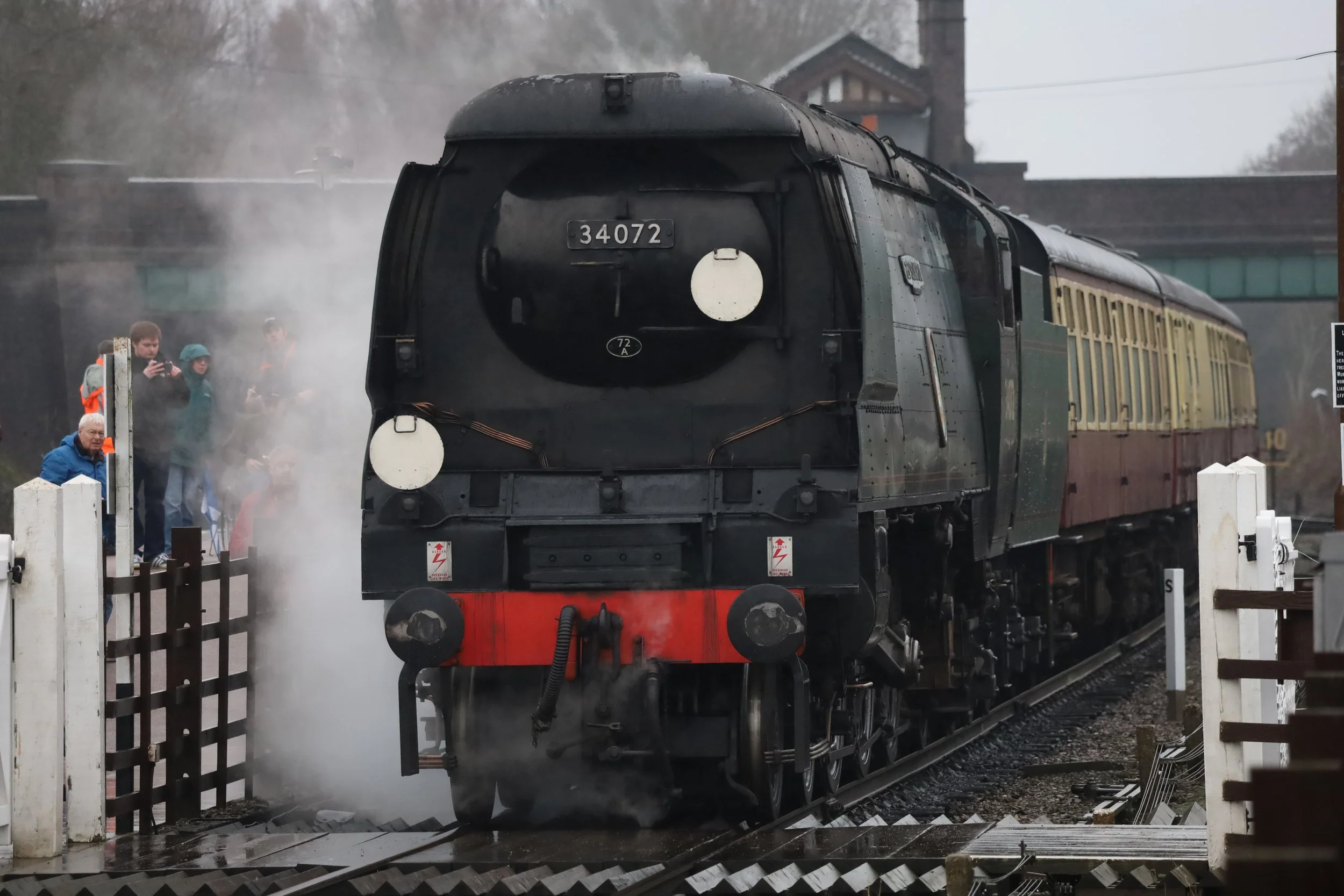 A vintage black steam locomotive with the number 34072 on the front, emitting steam, pulling passenger cars on a railway, with spectators observing nearby.
