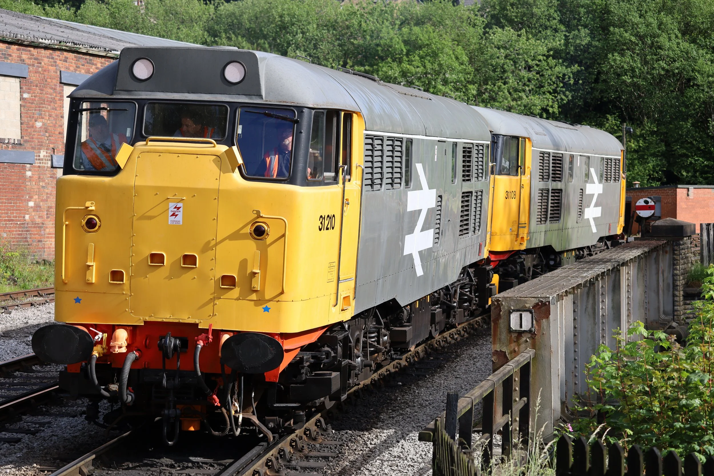 A yellow and gray train moving along a railway track with a brick building and green trees in the background.