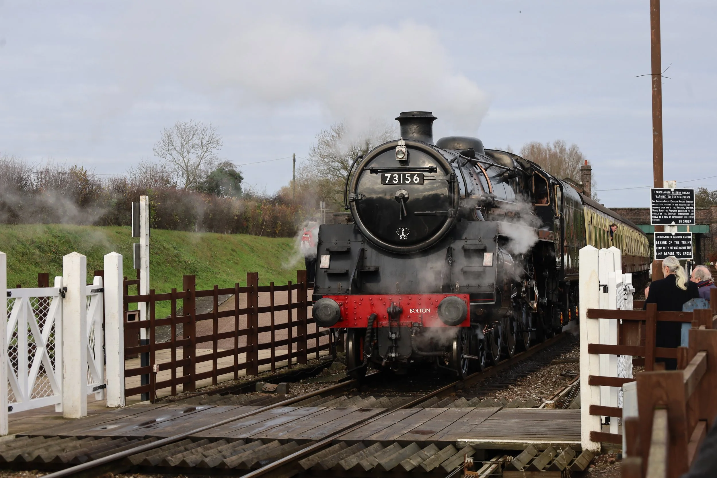 A vintage black steam locomotive train with the number 73156 is moving along the tracks, emitting steam and smoke, with a crowd of some people watching nearby.