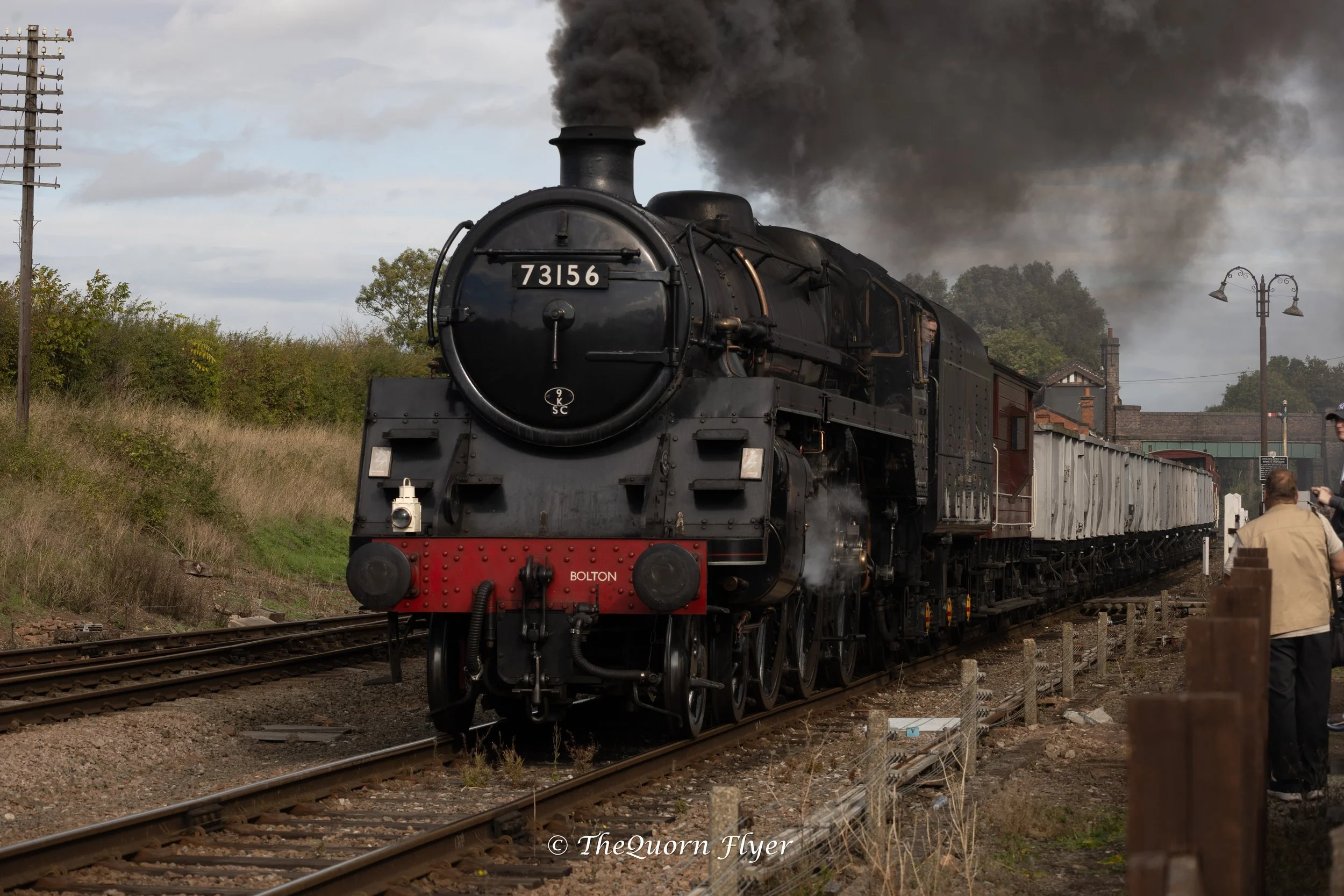 A black steam locomotive train with the number 73156 on the front, emitting black smoke, traveling on railway tracks with a surrounding rural landscape and a man taking a photo nearby.
