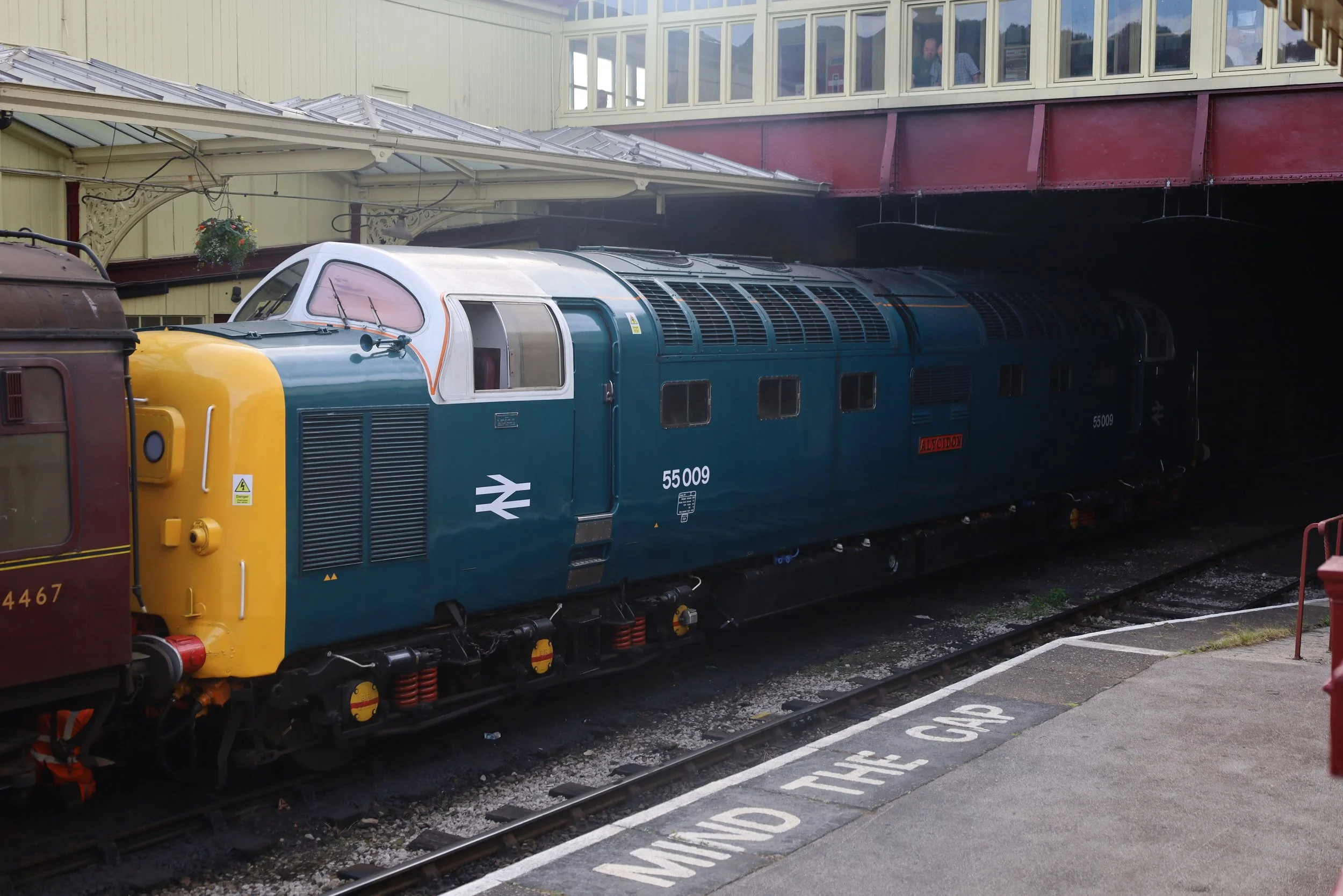Historical train engine painted blue and yellow at a station, with a maroon passenger car attached. The platform has a marked warning zone on the ground.