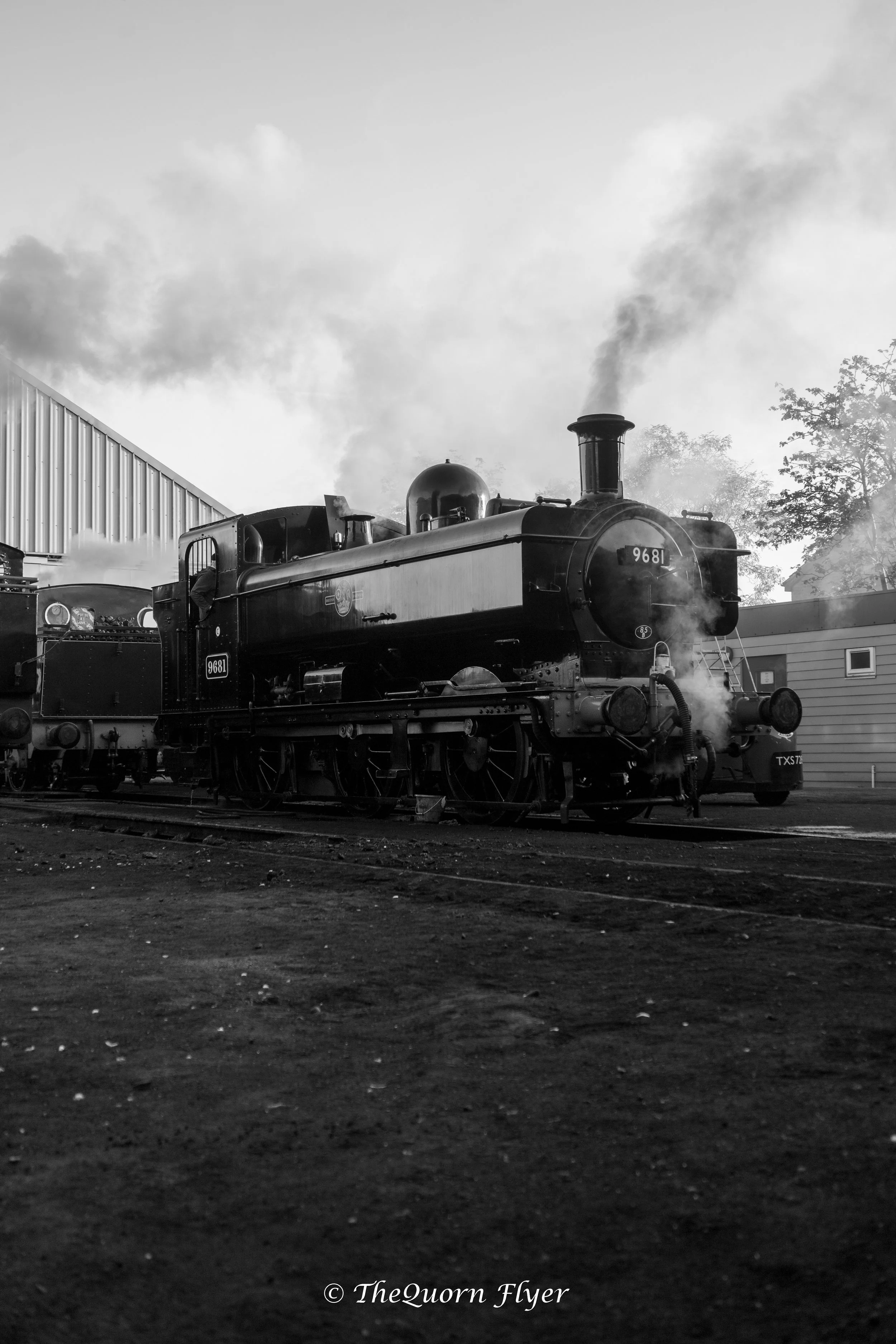 A vintage steam locomotive with the number 9681 on its front, emitting smoke as it sits on railway tracks near a building and trees, photographed in black and white.