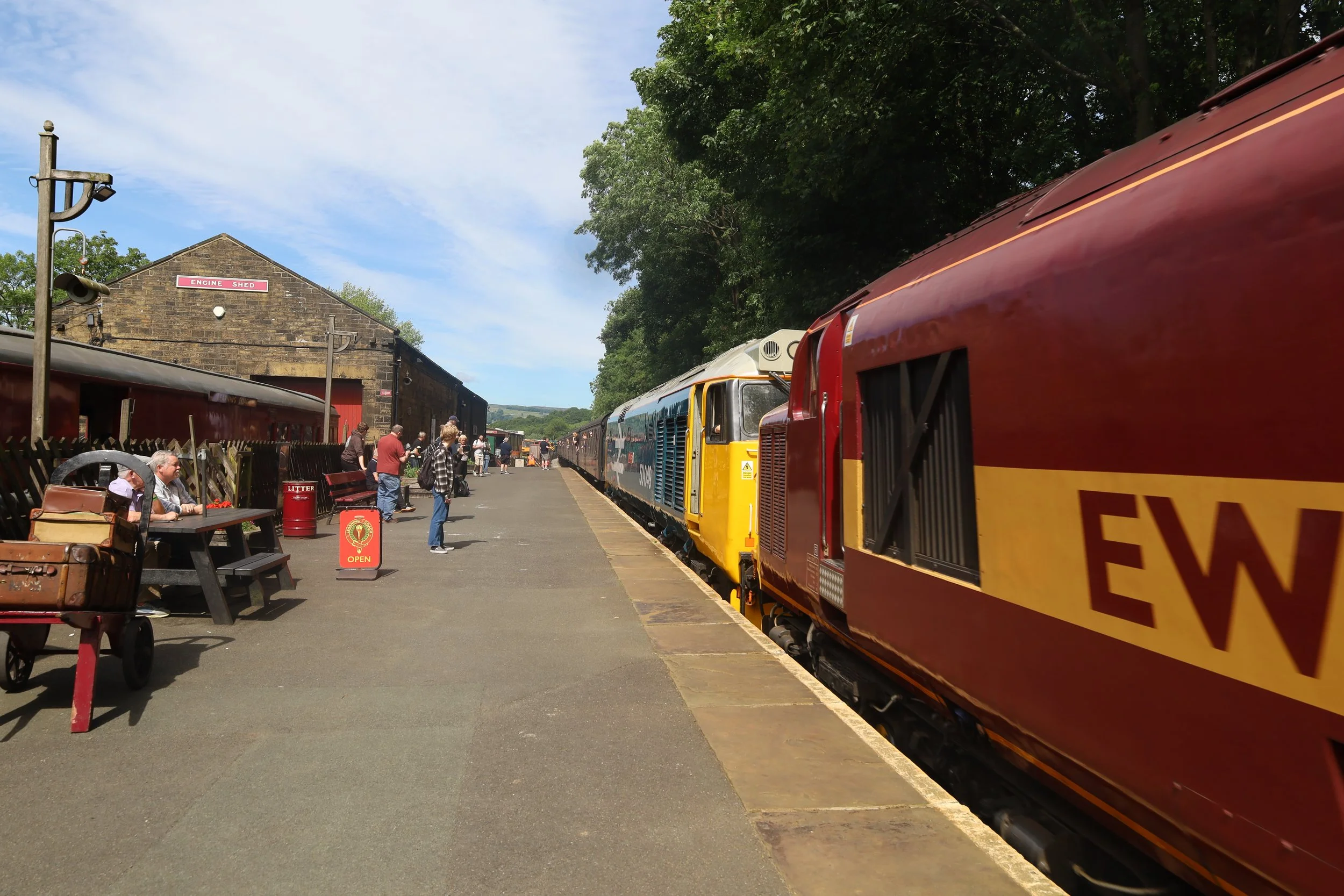 A train station with multiple colorful train cars parked along the platform. People are sitting on benches and walking around. There is a brick building labeled 'Engine Shed' in the background, with trees and a blue sky with some clouds overhead.